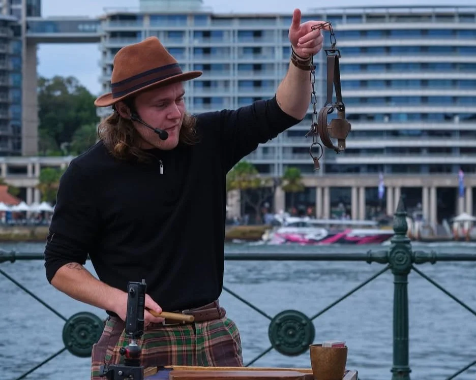 A magician in a brown fedora hat and black shirt holding a chain with a pair of handcuffs on it, possibly performing a street act or demonstration near a waterfront with boats and a cityscape in the background.
