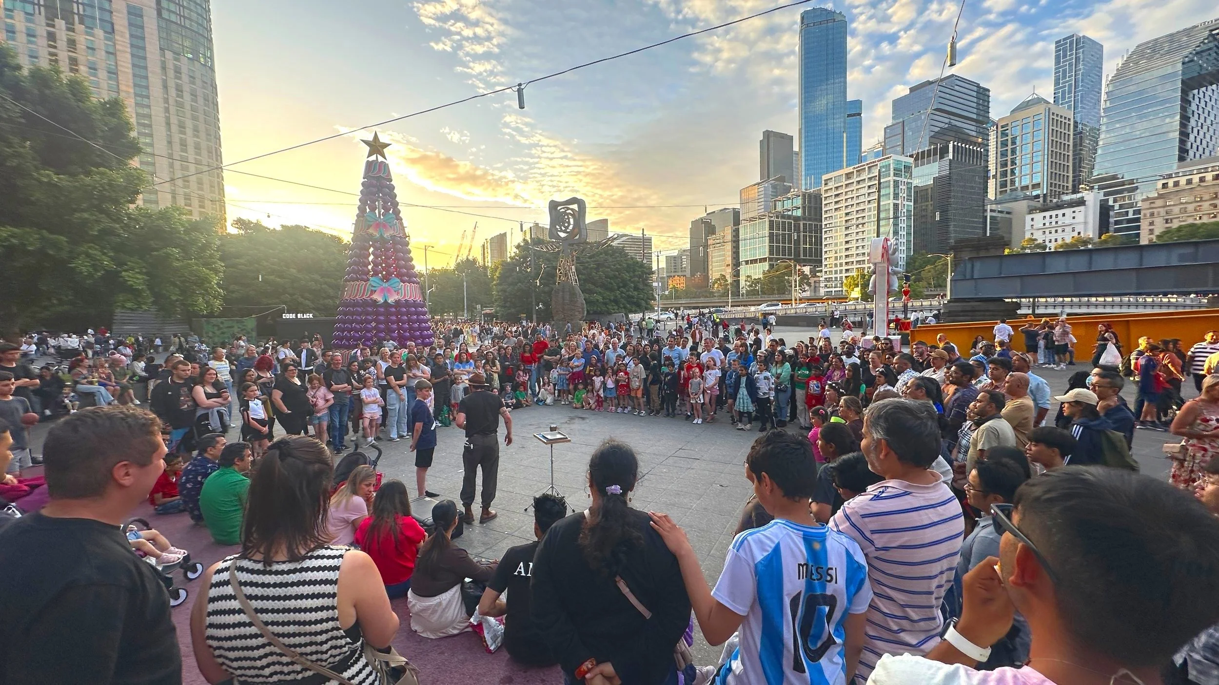 A large crowd of people gathered outdoors in front of a city skyline at sunset, with holiday-themed decorations and a Christmas tree in the background to watch a street magic show