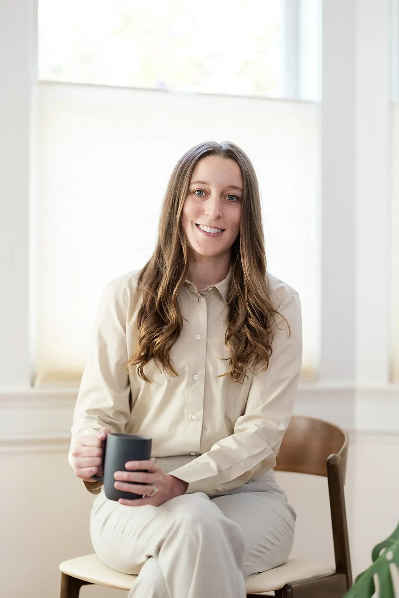 Sexual assault therapist Michele Root, LCSW smiles warmly, holding a dark grey mug. She has long brown wavy hair and wears a beige button-down work shirt.