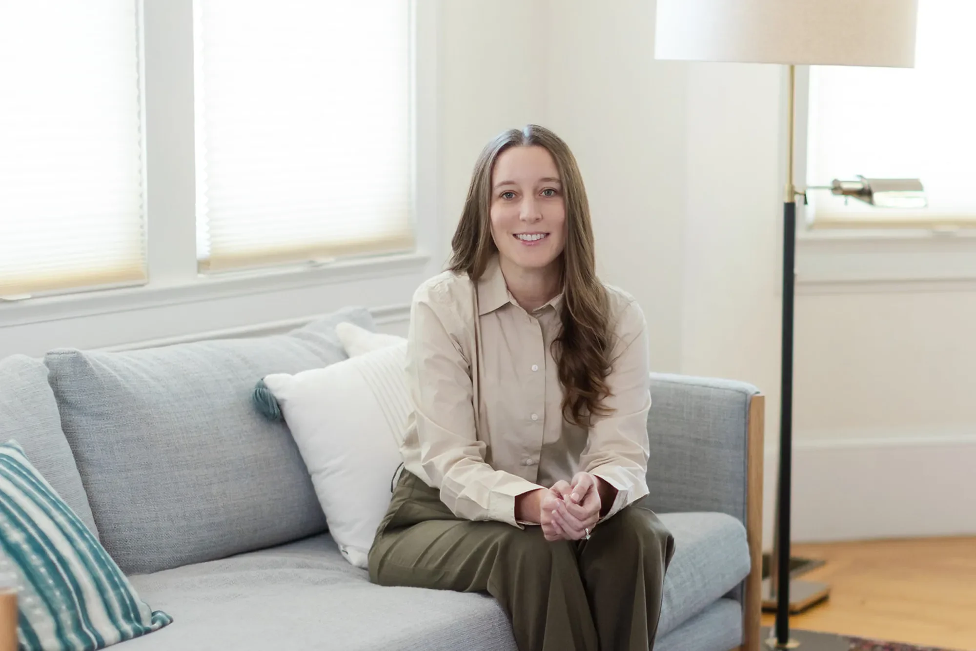Domestic violence therapist Michele Root, LCSW sits on a grey couch with her hands together on her lap in a brightly lit room. She smiles warmly, wears a beige button-down shirt, and has long brown wavy hair.