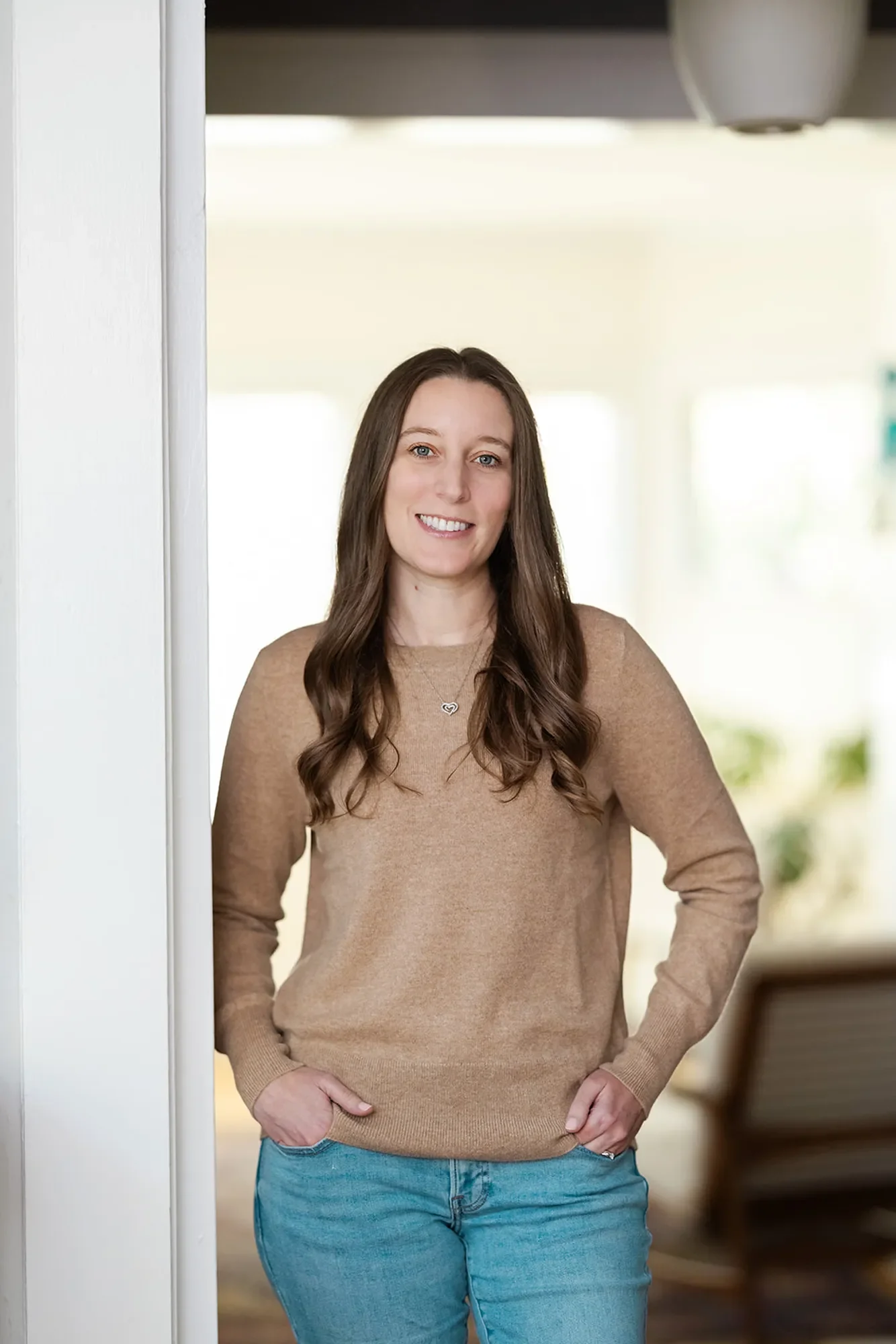 Abuse therapist Michele Root, LCSW smiles warmly with her hands in her pockets in a well-lit room. She has long, brown, wavy hair and is wearing a beige sweater.