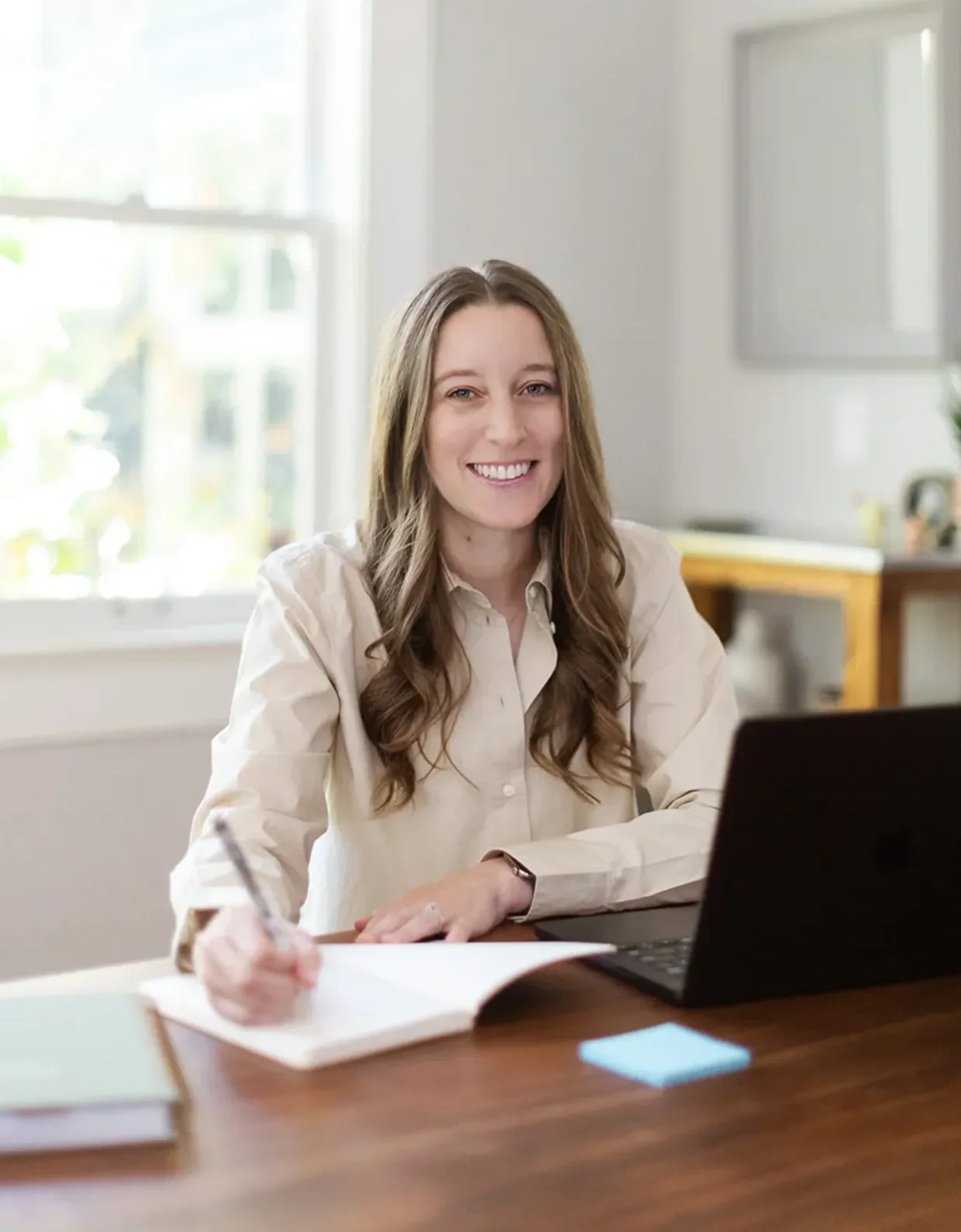 Michele Root, LCSW smiles while sitting at a desk with a laptop, writing in a notebook. Sunlight comes through a window behind her, and the room has a bright, modern look.