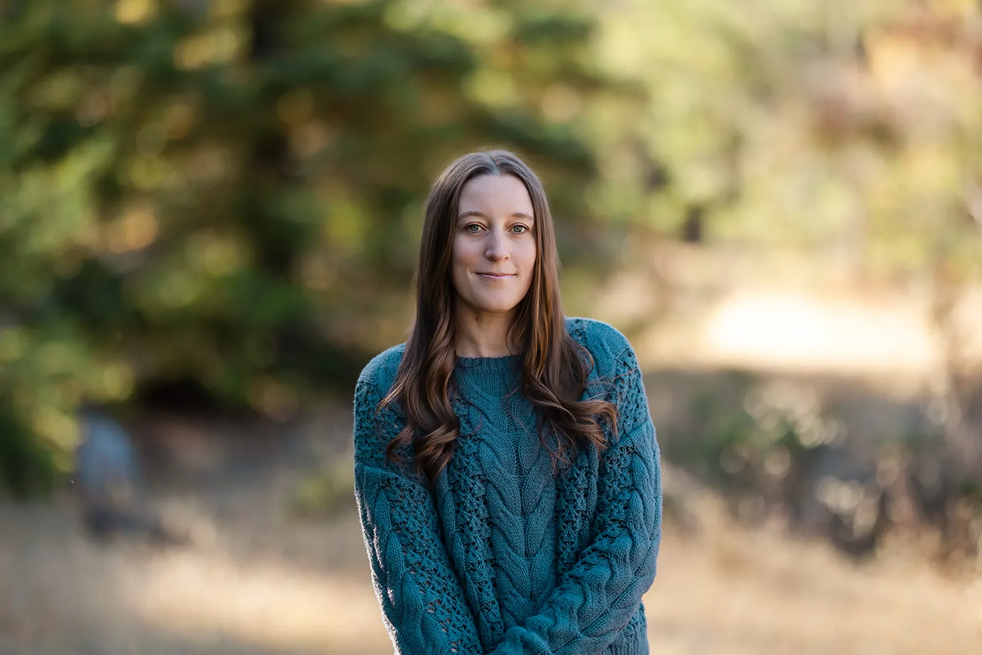 First responder therapist Michele Root, LCSW wears a teal cableknit sweater, and stands smiling gently in a grassy field with trees in the background.