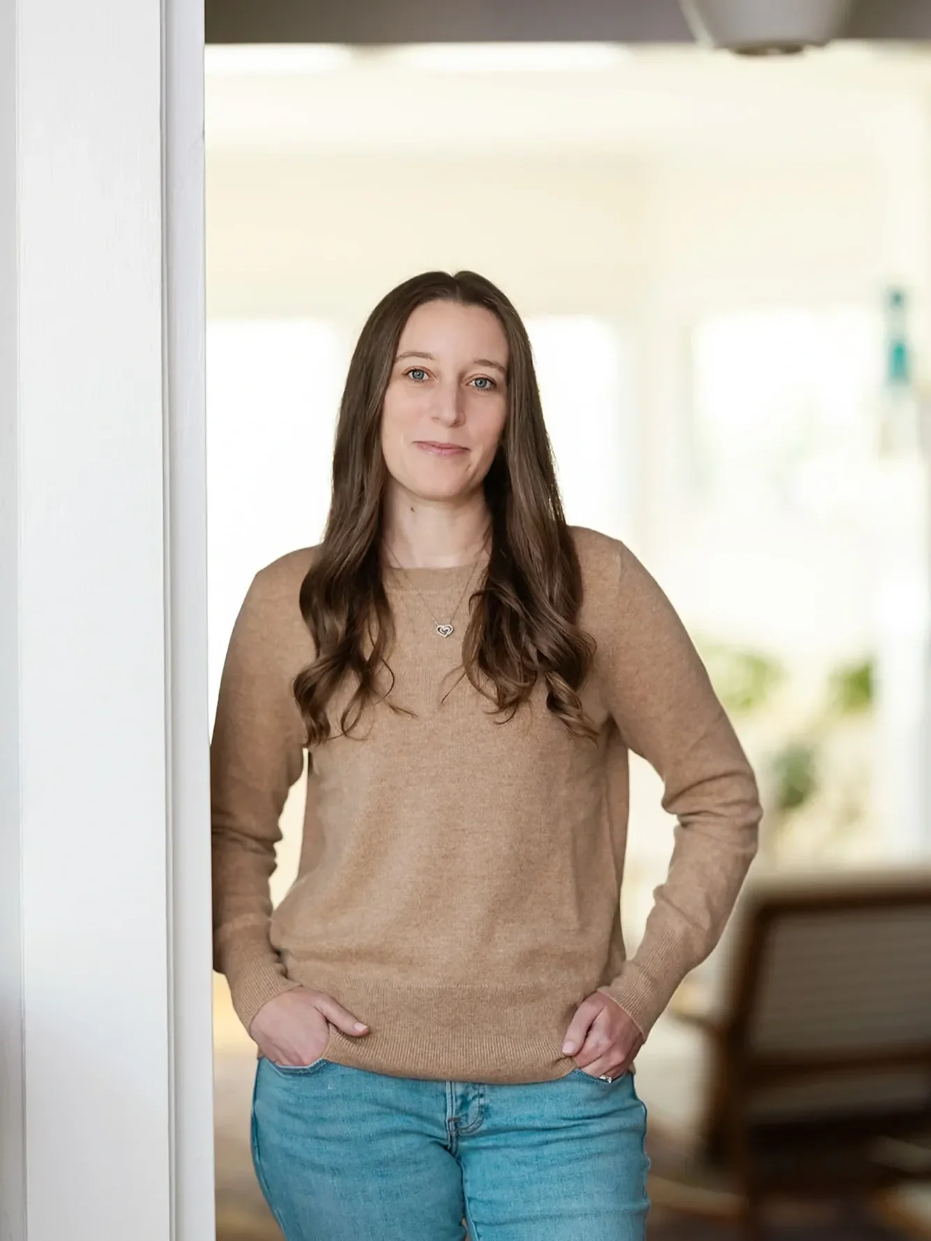 Michele Root, LCSW is wearing a light brown sweater and blue jeans, stands indoors with her hands in her pockets, smiling softly at the camera.