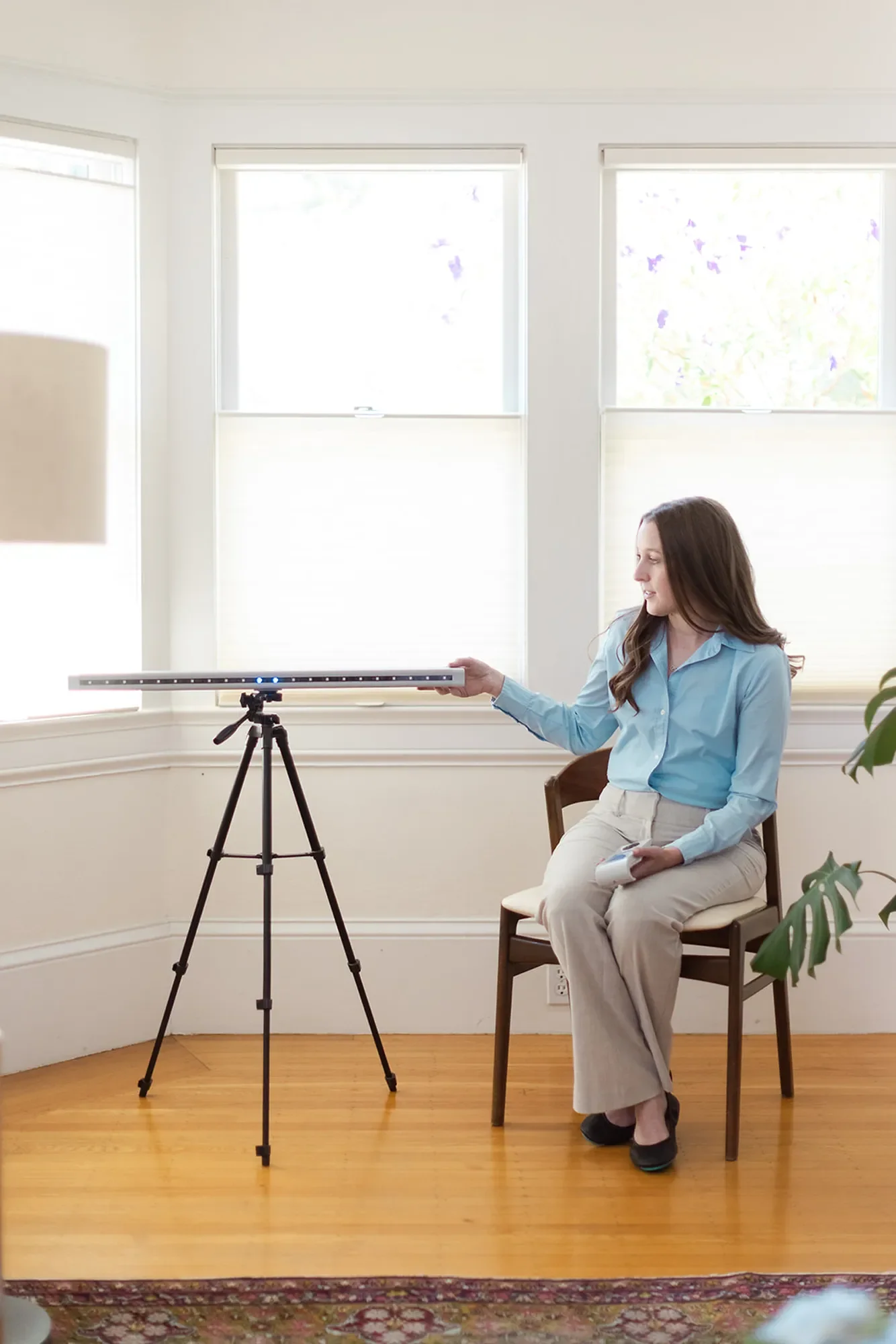 Michele Root, LCSW sits on a chair and adjusts an EMDR bilateral stimulation device that’s mounted on a tripod in a bright room with large windows.