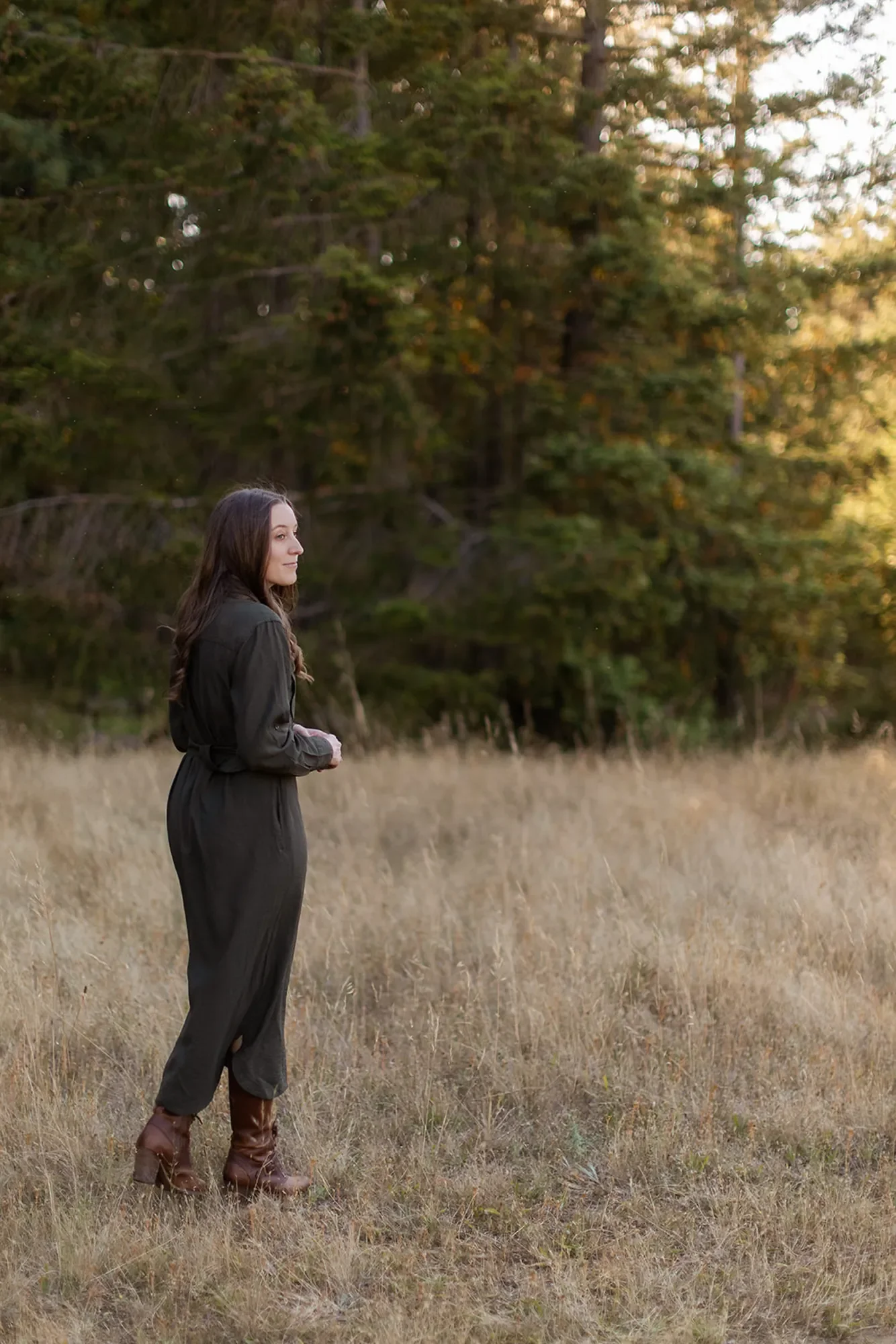 Trauma therapist Michele Root, LCSW is wearing a long dark dress and brown boots, and stands in a grassy field, facing right, with tall green pine trees in the background.