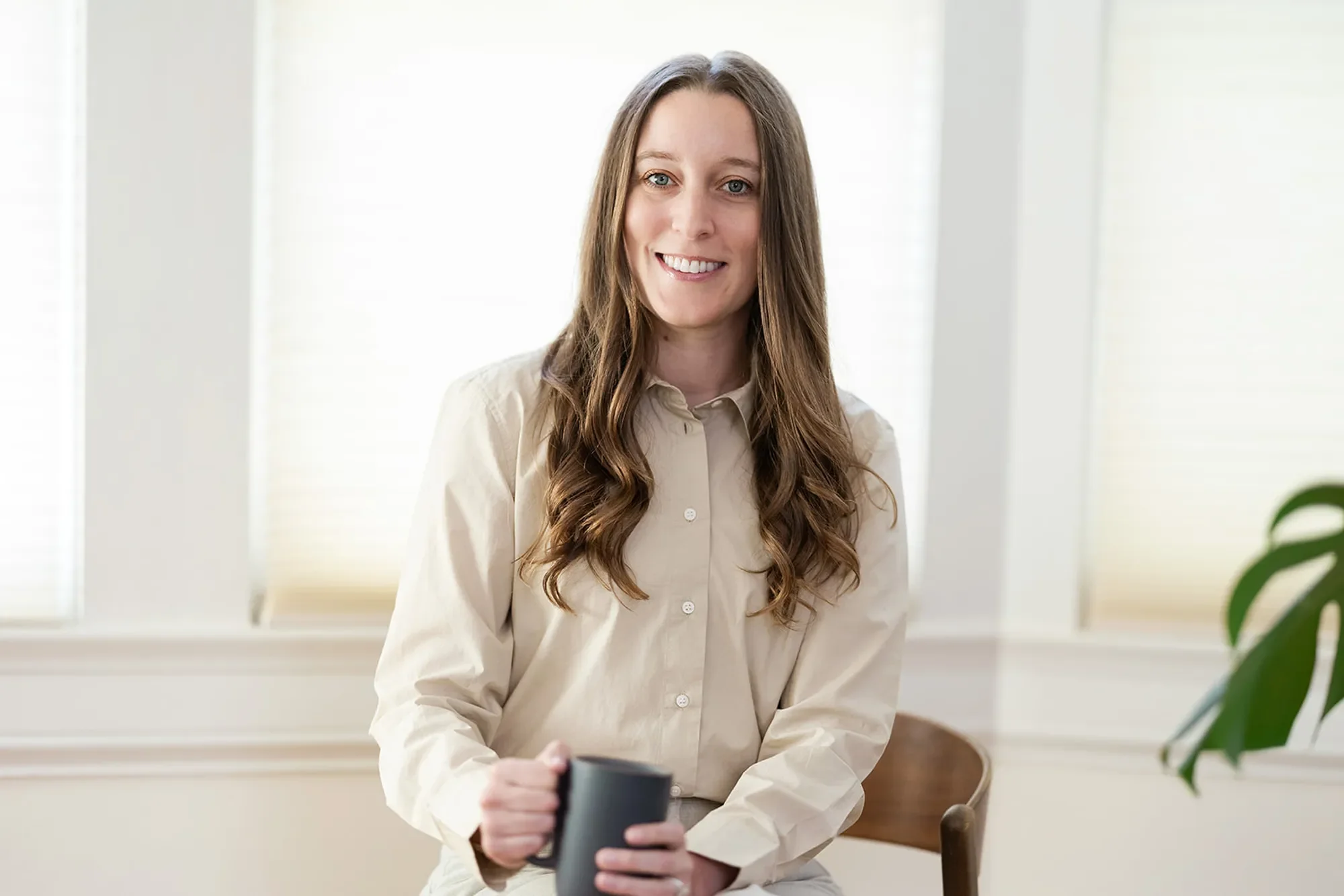 Therapist Michele Root, LCSW smiles warmly, holding a dark grey mug. She has long brown wavy hair and wears a beige button-down work shirt.