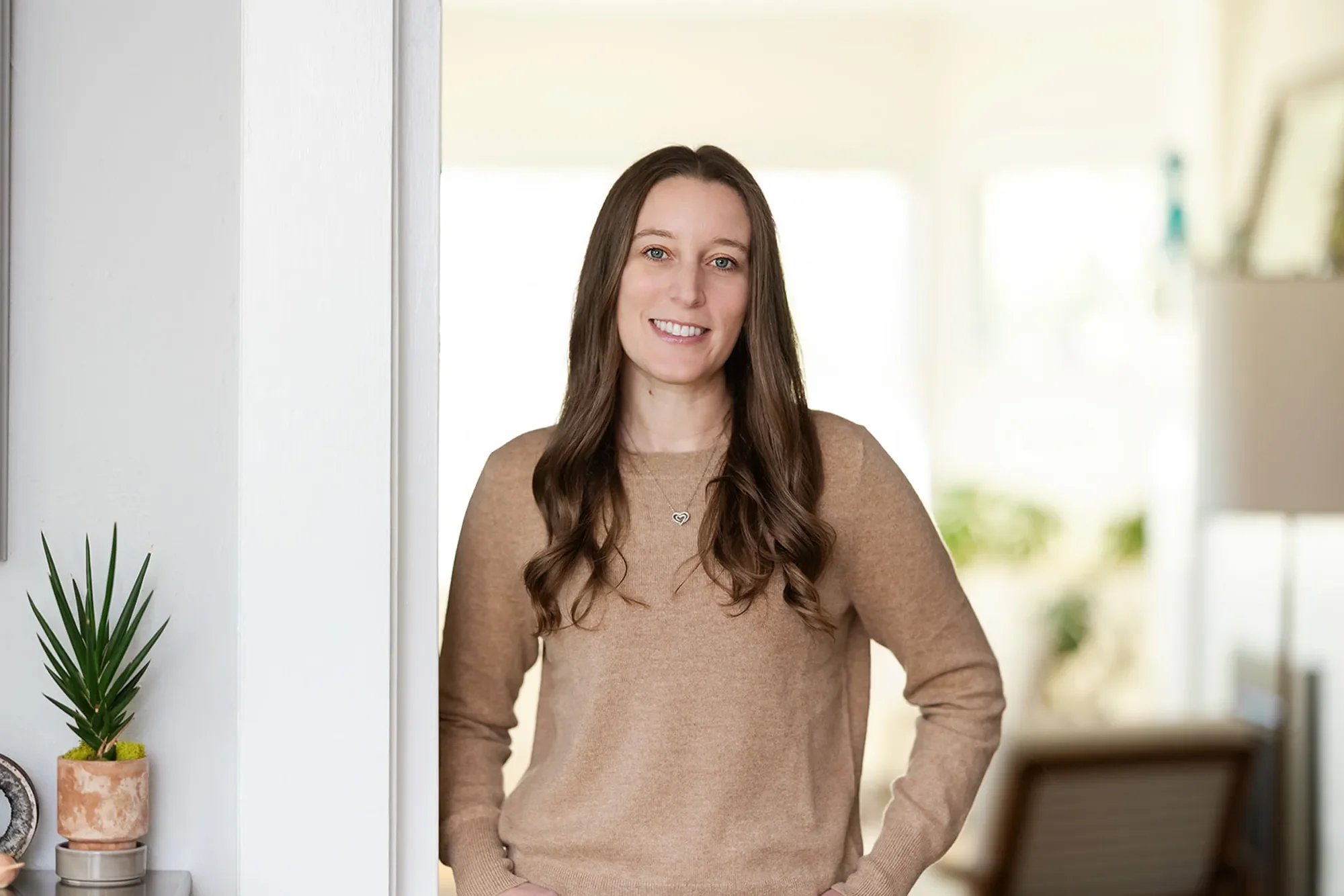 Therapist for first responders Michele Root, LCSW smiles warmly with her hands in her pockets in a well-lit room. She has long, brown, wavy hair and is wearing a beige sweater.