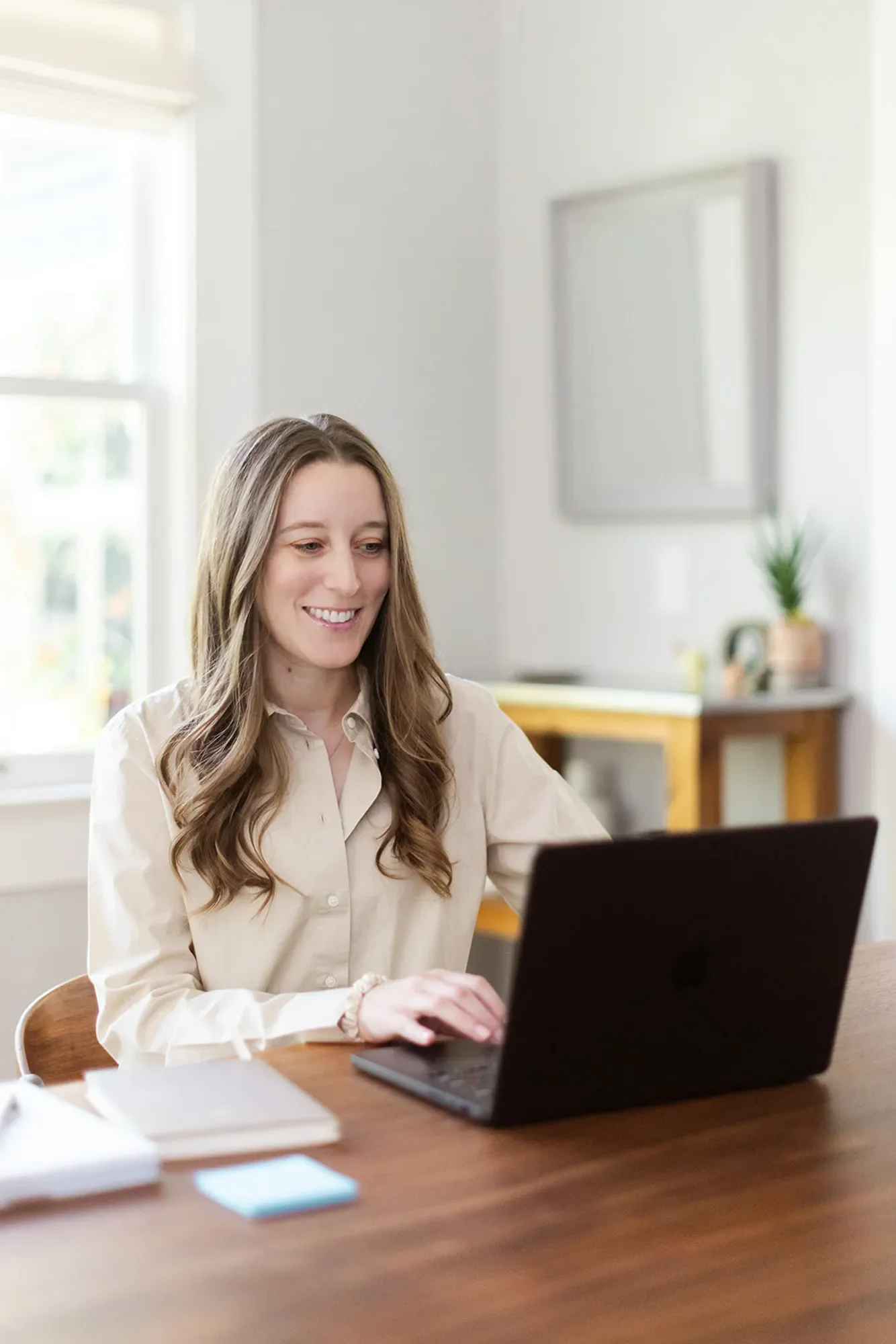 Therapist Michele Root, LCSW smiles as she types on a laptop. There is a console table with a plant and mirror in the background, in a brightly lit room.