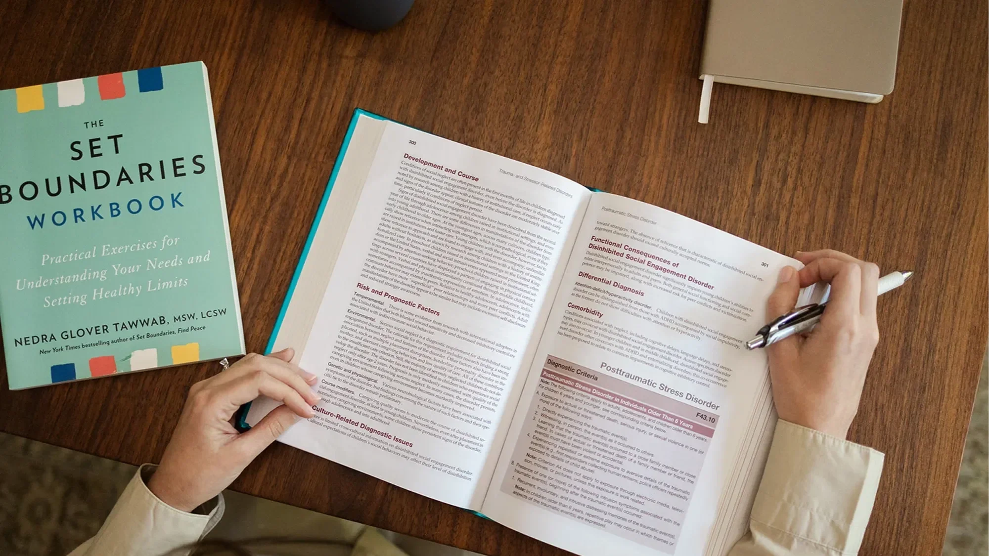 Michele Root, LCSW holds open a mental health textbook next to The Set Boundaries Workbook on a wooden table. The pages discuss psychological conditions with highlighted sections.