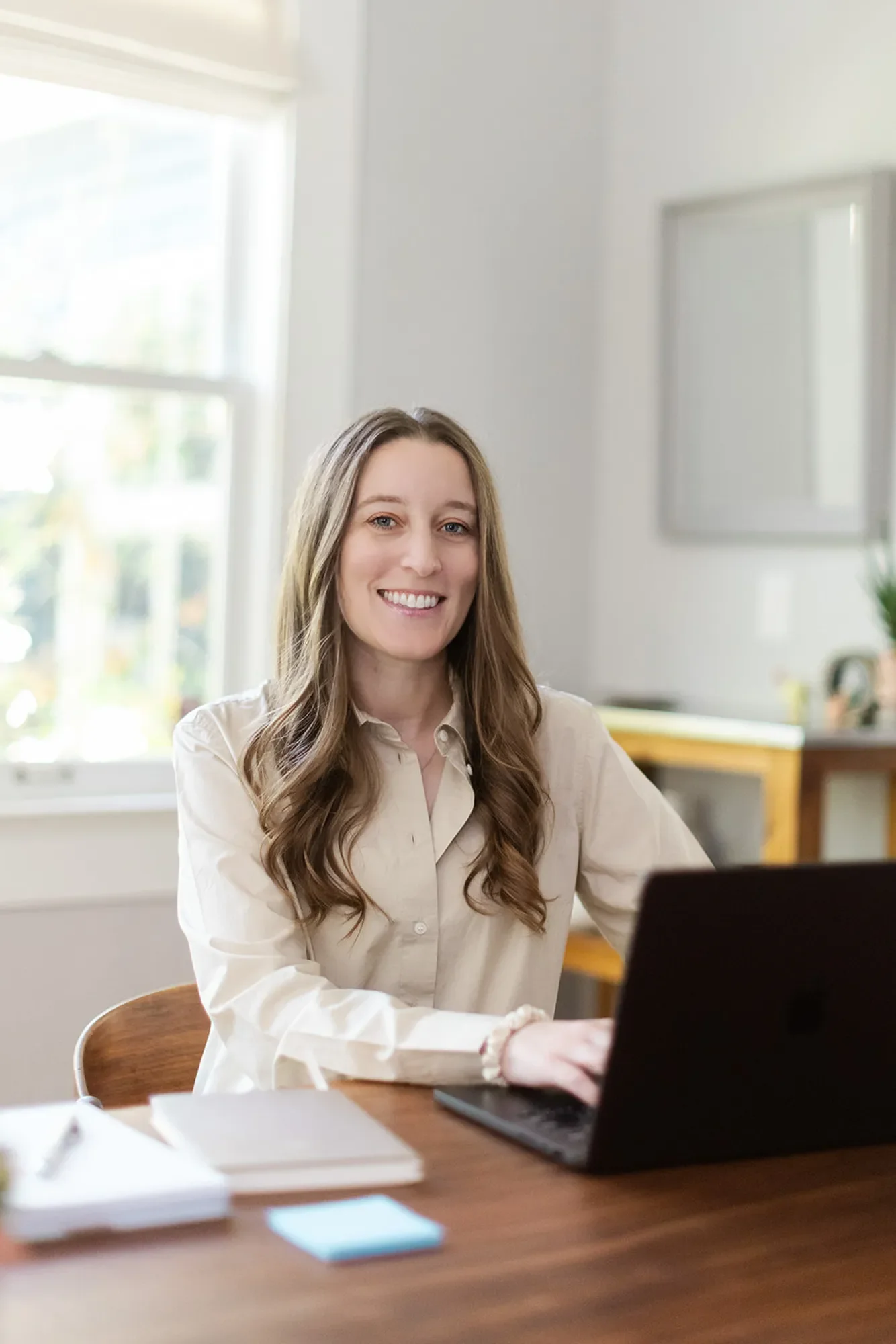 EMDR therapist Michele Root, LCSW has long brown hair, wears a beige dress shirt, and sits at a desk using a laptop and smiling. There is a notebook and pen on the desk, with a bright window and shelves in the background.