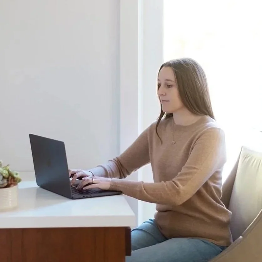 Michele Root, LCSW sits at a desk using a laptop in a bright room with natural light coming through a nearby window.