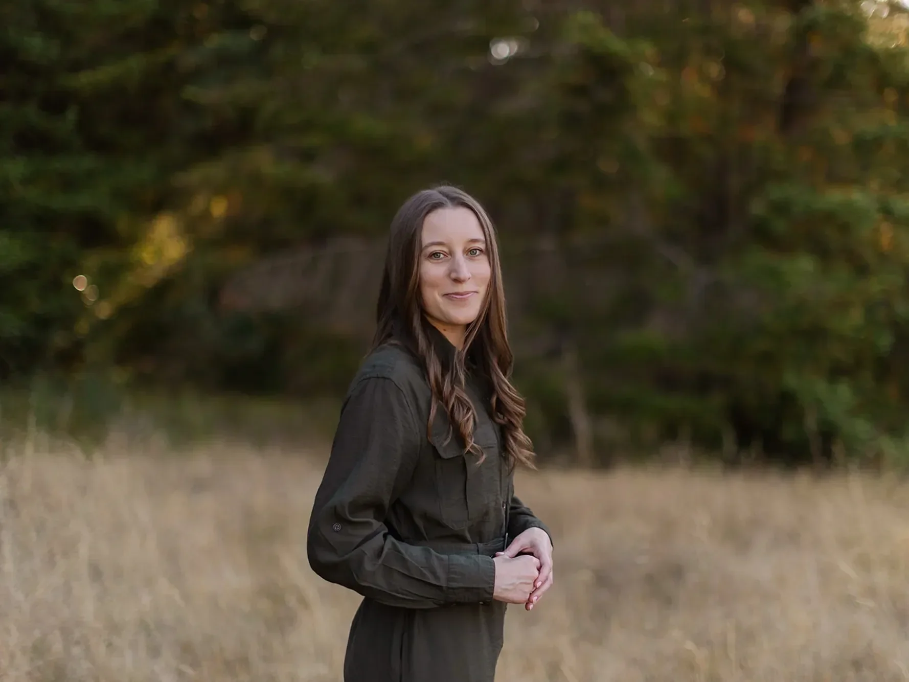 Therapist Michele Root, LCSW has long brown hair, is wearing a dark green long dress, and stands in a dry grassy field with green trees in the background, smiling slightly at the camera.