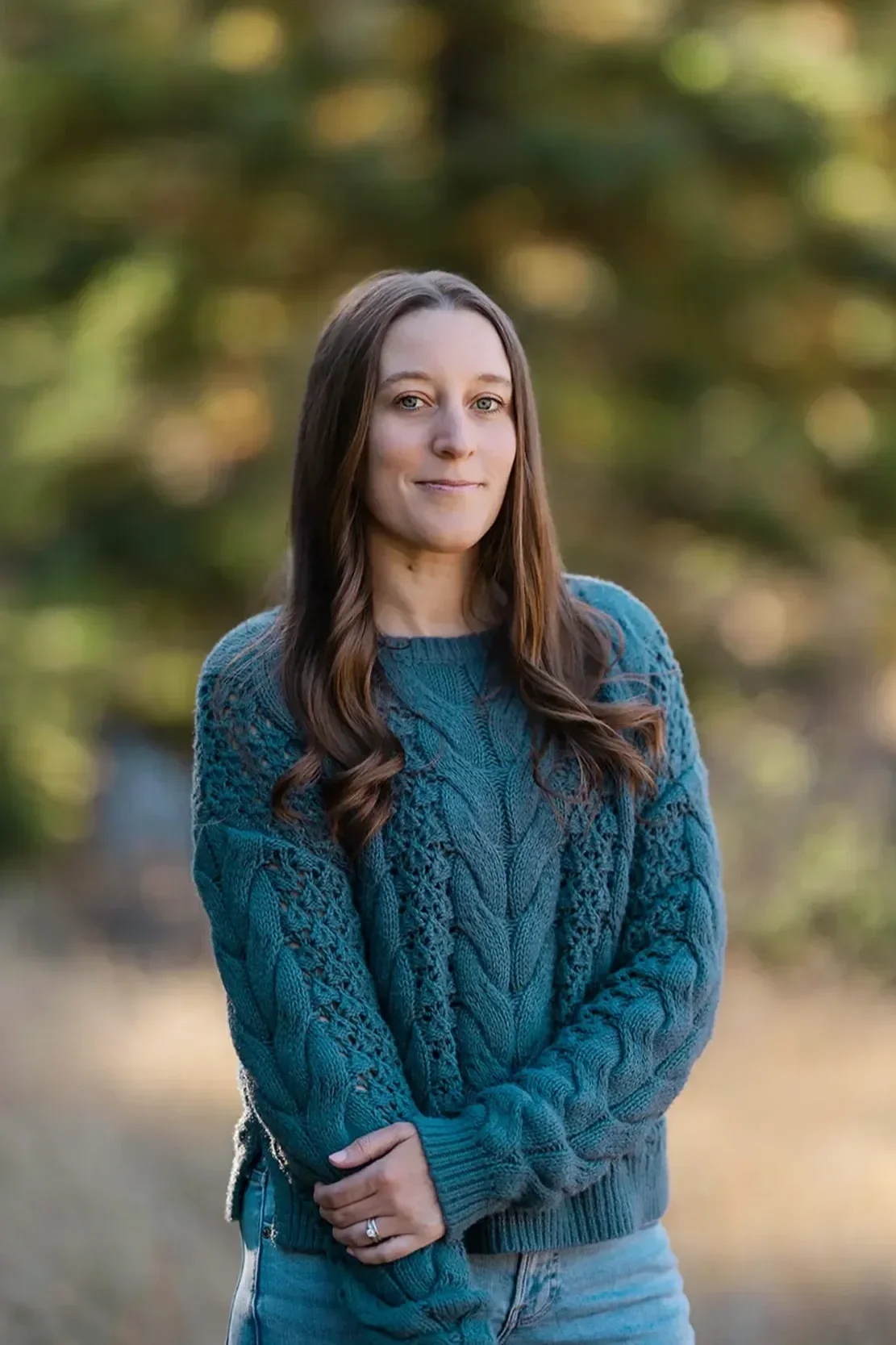 Michele Root, LCSW has long brown hair, wears a teal knit sweater and jeans, stands outdoors with her arms crossed. The background is blurred with green foliage and sunlight.