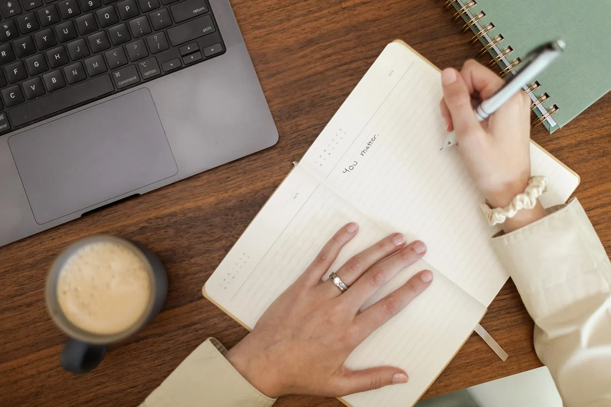 Michele Root, LCSW writes “you matter” in a notebook on a wooden desk, beside a laptop, a spiral notebook, and a coffee cup.