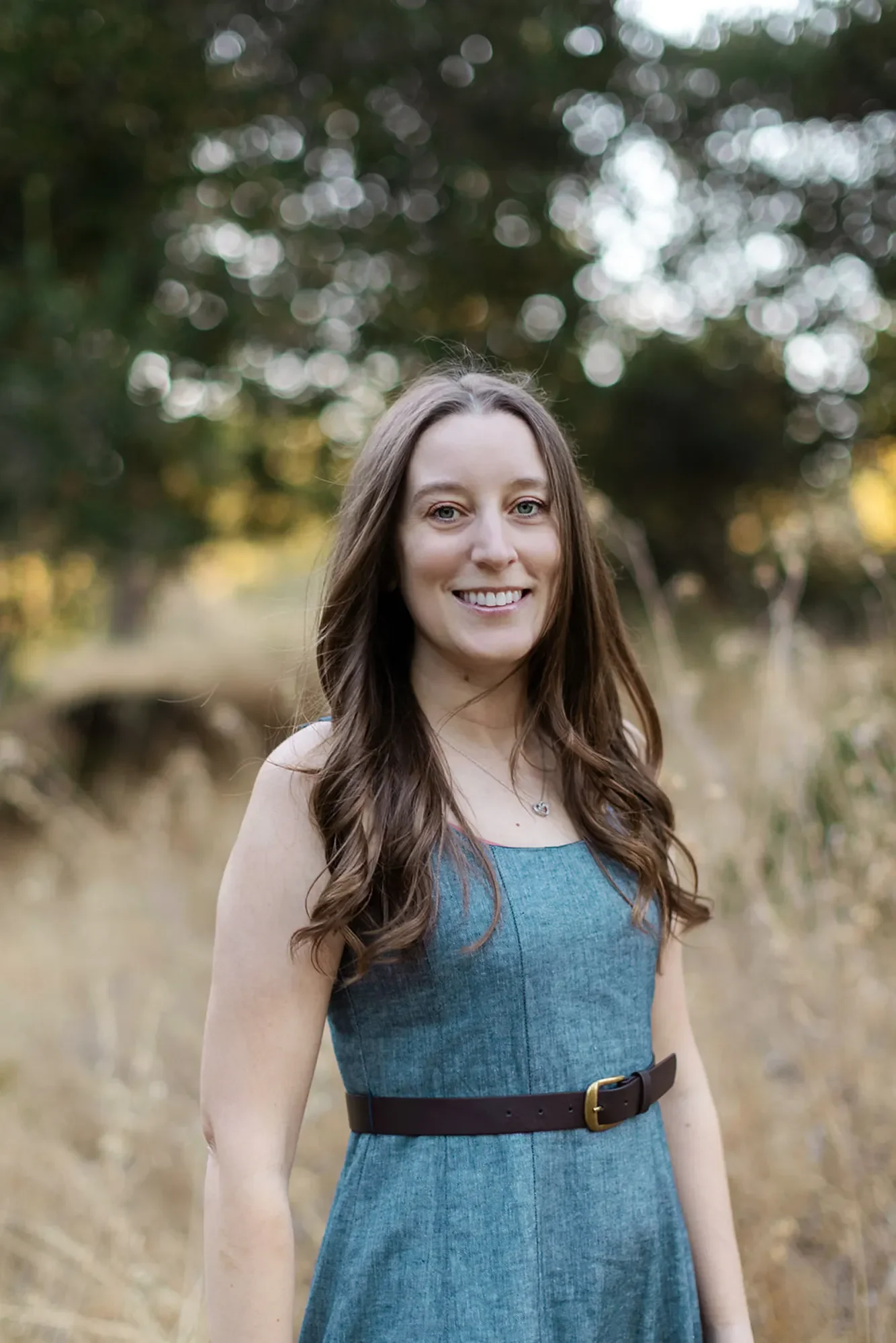 Therapist Michele Root, LCSW stands smiling in a field with tall, dry grass, with trees in the background. She has long, brown wavy hair and wears a blue dress with a brown belt.