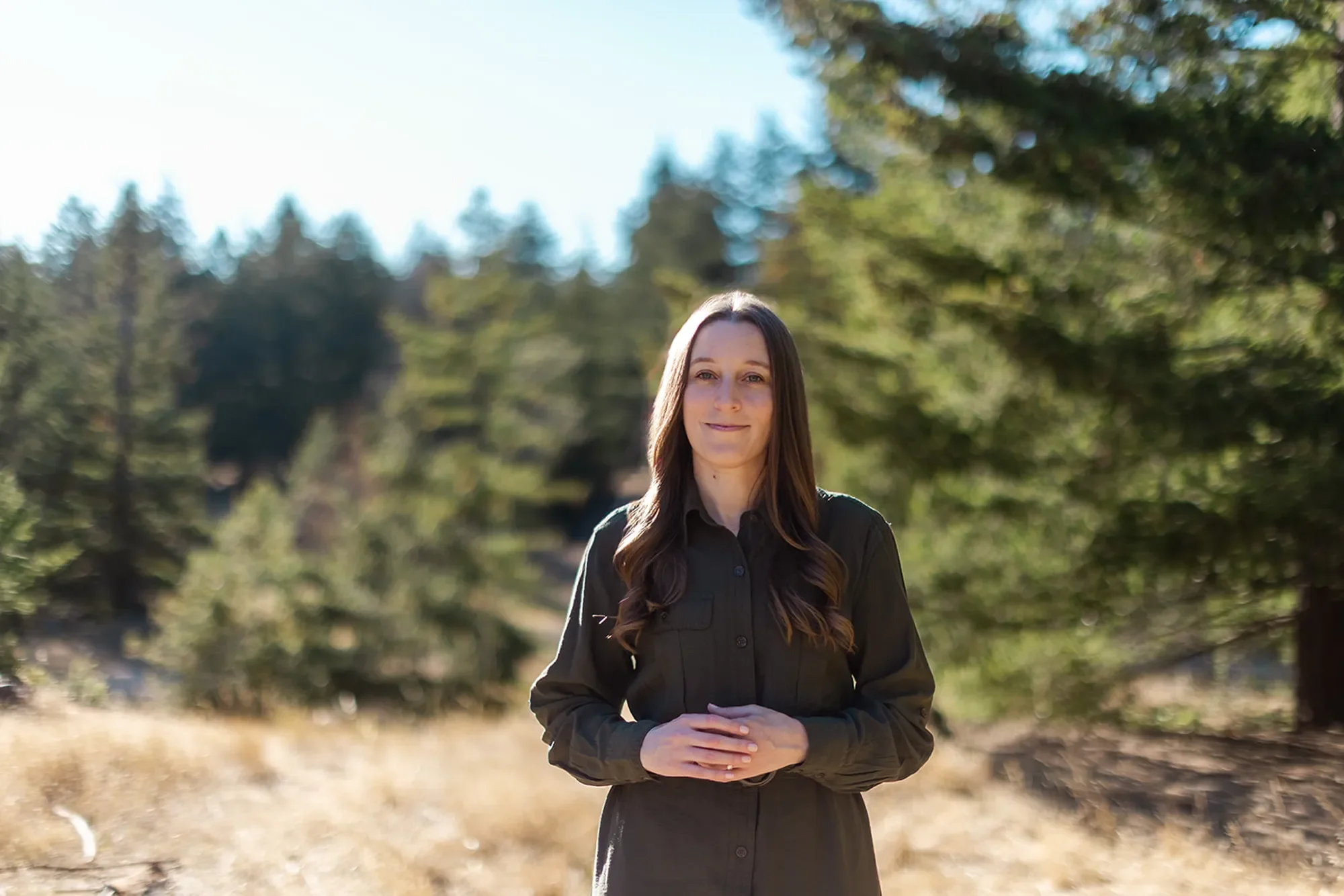 Sexual abuse therapist Michele Root, LCSW stands in a long dark dress in a sunlit field with tall grass, surrounded by trees and a forest background under a clear sky. Her hands are clasped in front of her as she smiles softly.