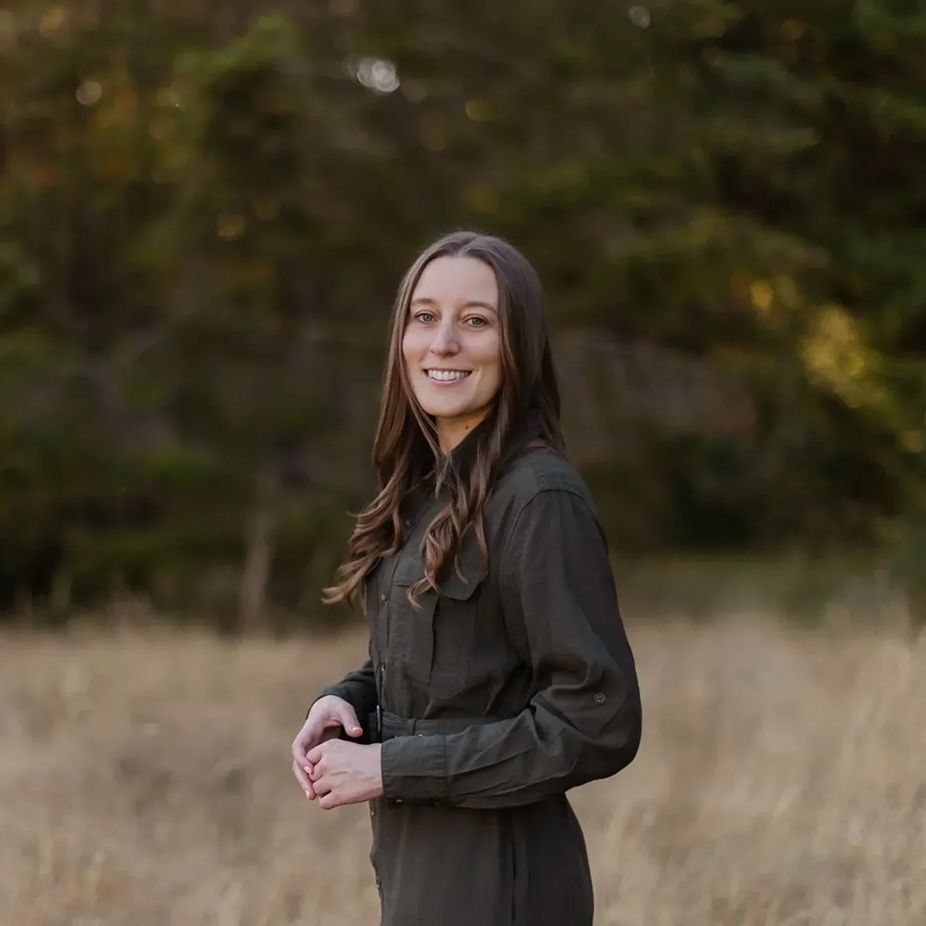 Childhood trauma therapist Michele Root, LCSW stands in a long dark dress in a sunlit field with tall grass, surrounded by trees. Her hands are held in front of her as she smiles softly.