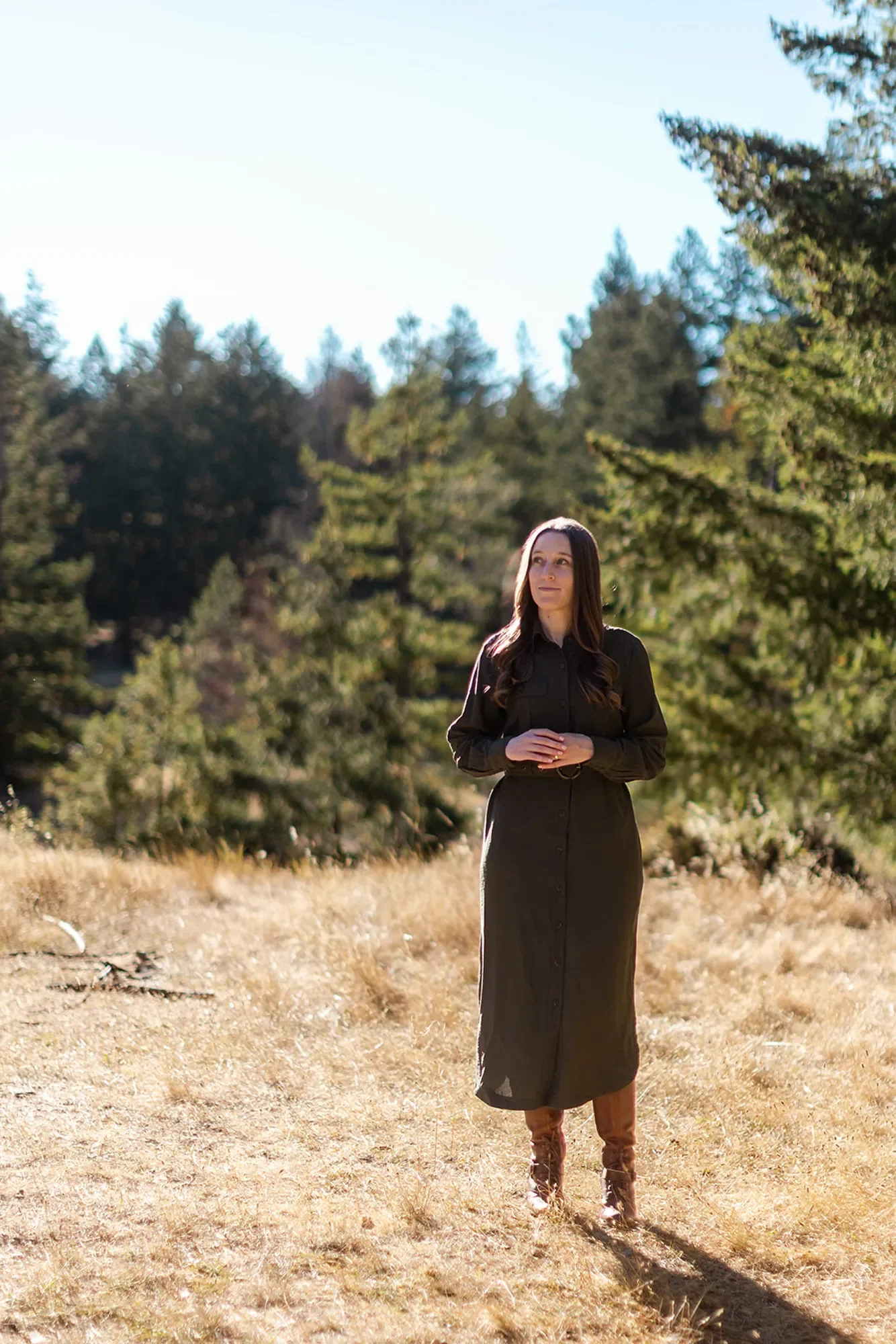 EMDR trauma therapist Michele Root stands in a long dark dress in a sunlit field with tall grass, surrounded by trees and a forest background under a clear sky.