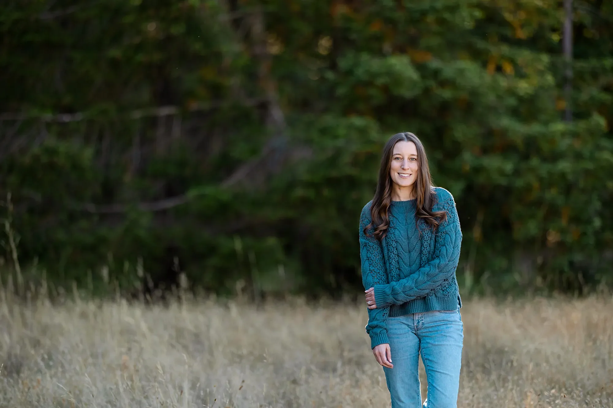 Michele Root, LCSW is wearing a teal sweater and jeans, stands smiling in a grassy field with trees in the background.