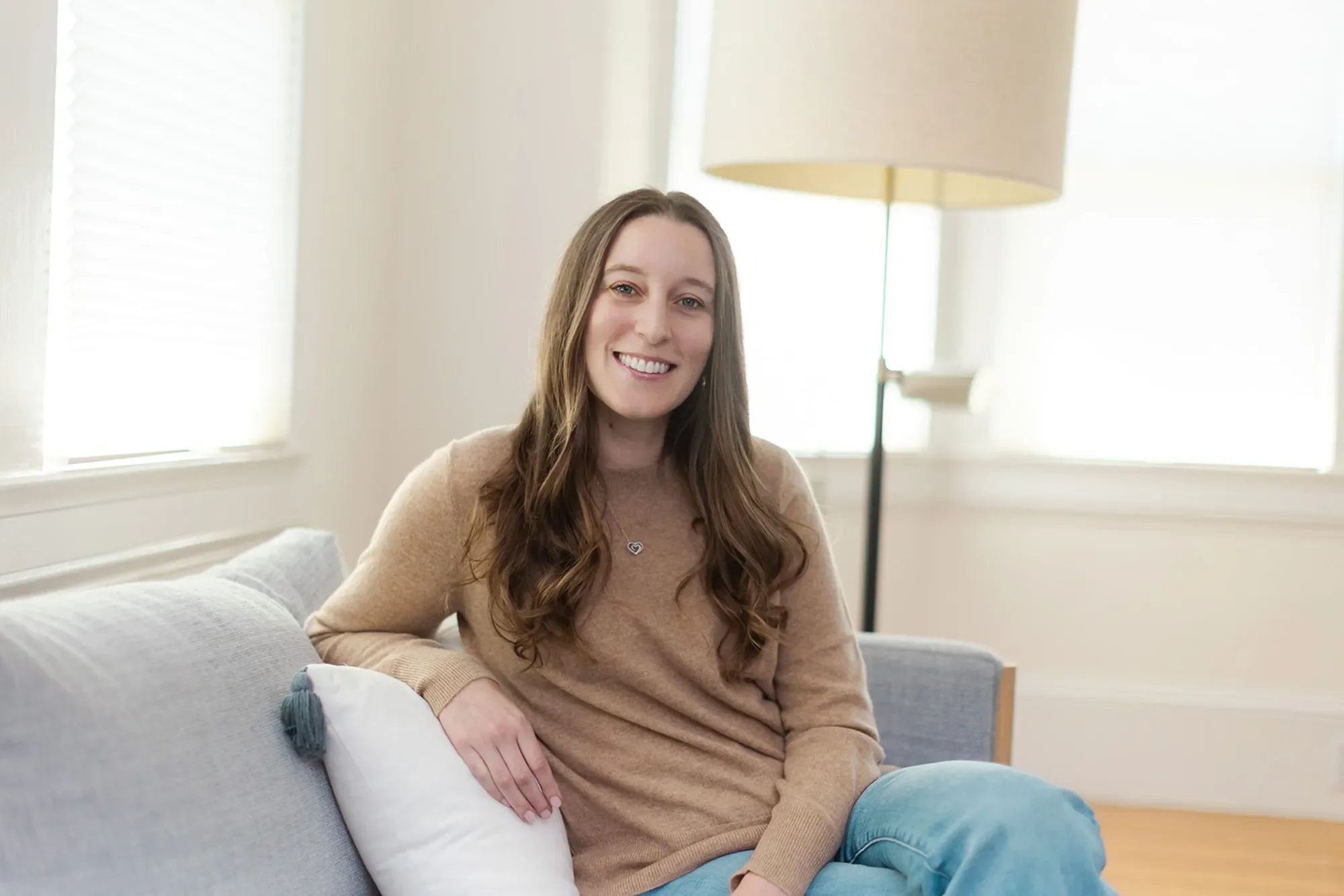 Therapist Michele Root, LCSW sits cross-legged on a grey couch in a bright room, smiling warmly. She has long, wavy brown hair and wears a camel-colored sweater and jeans.