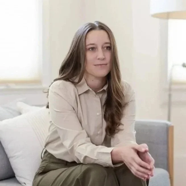 Michele Root, LCSW has long brown hair and wears a beige dress shirt. She sits on a couch as she listens intently to a person who is off-camera.