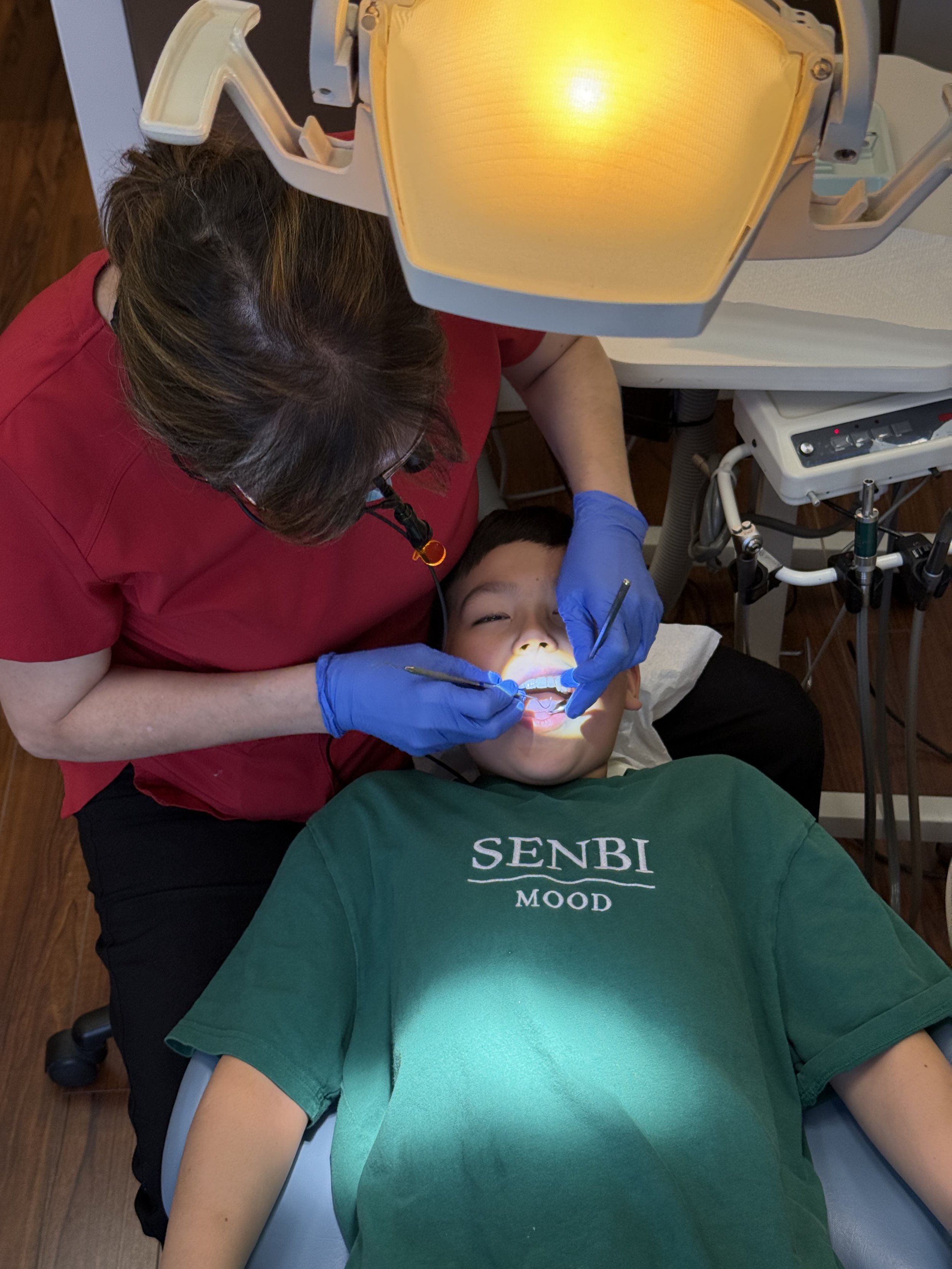 Dentist performing a check-up on a young patient lying in a dental chair, under a dental light, wearing a green shirt.