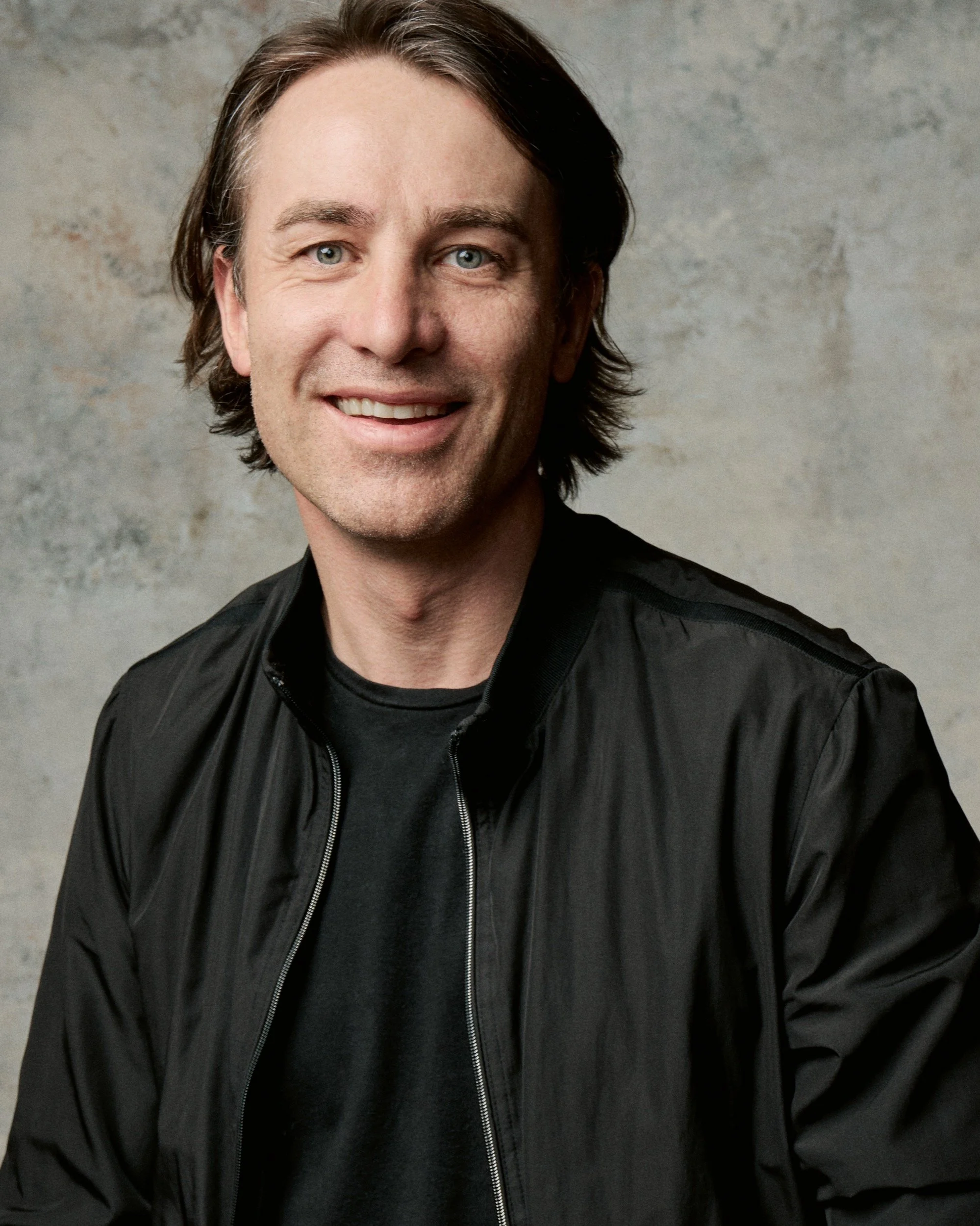 Close-up portrait of co-founder Andrew J Morley, a smiling middle-aged man with light skin, blue eyes, dark brown hair, and a slight beard, wearing a black jacket and black shirt, against a light gray textured background.