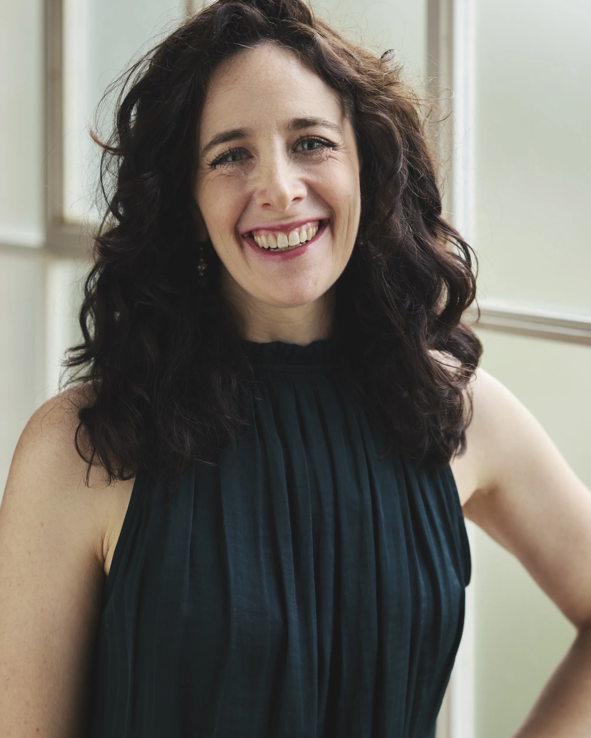 Close-up portrait of co-founder Kimberley Duband, a smiling woman with dark, wavy hair pictured indoors by a window, wearing a dark pleated top.