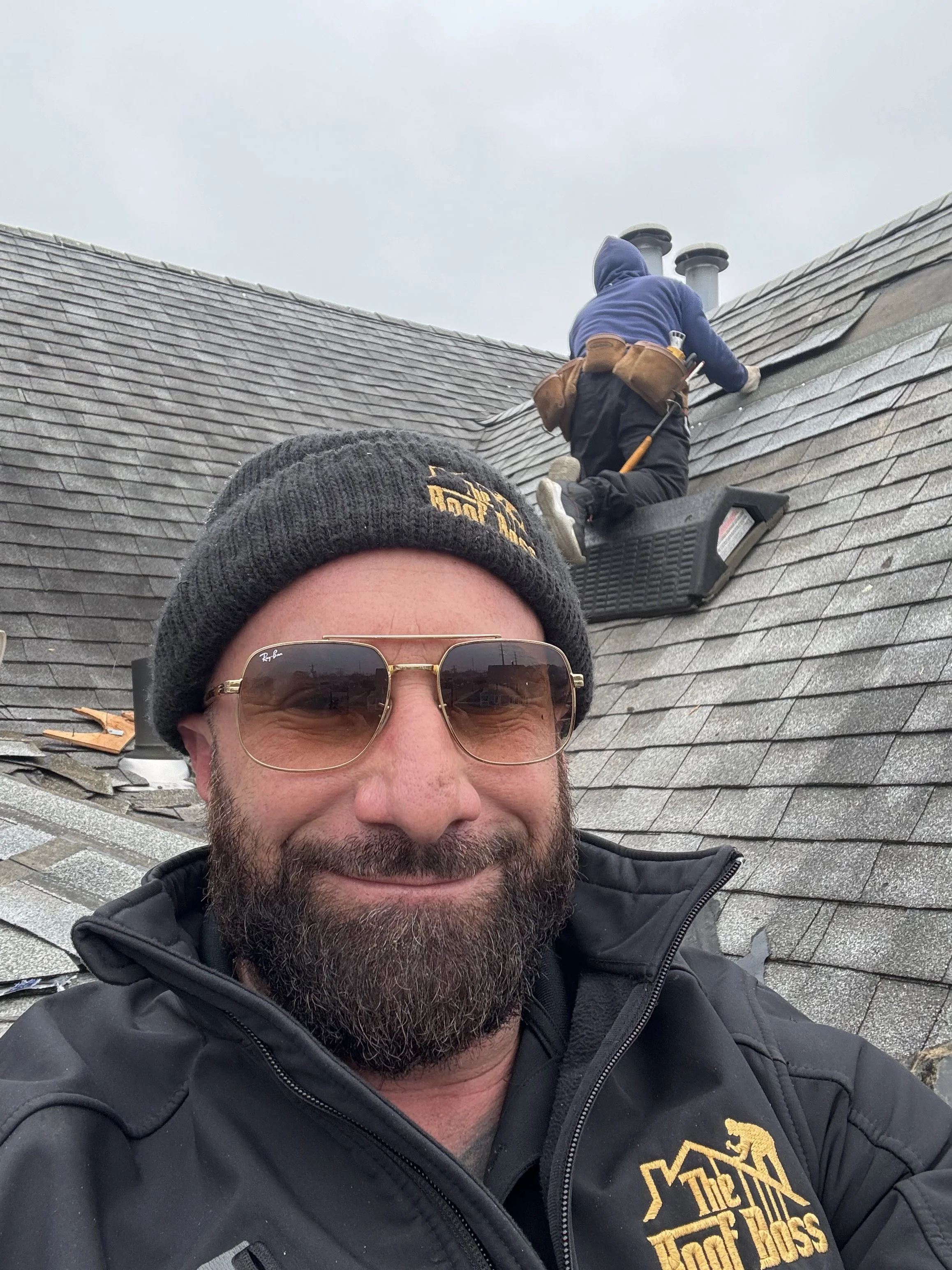 A man smiling wearing sunglasses, a grey beanie, and a black jacket, standing on a roof while two workers install or repair shingles in a cloudy sky background.