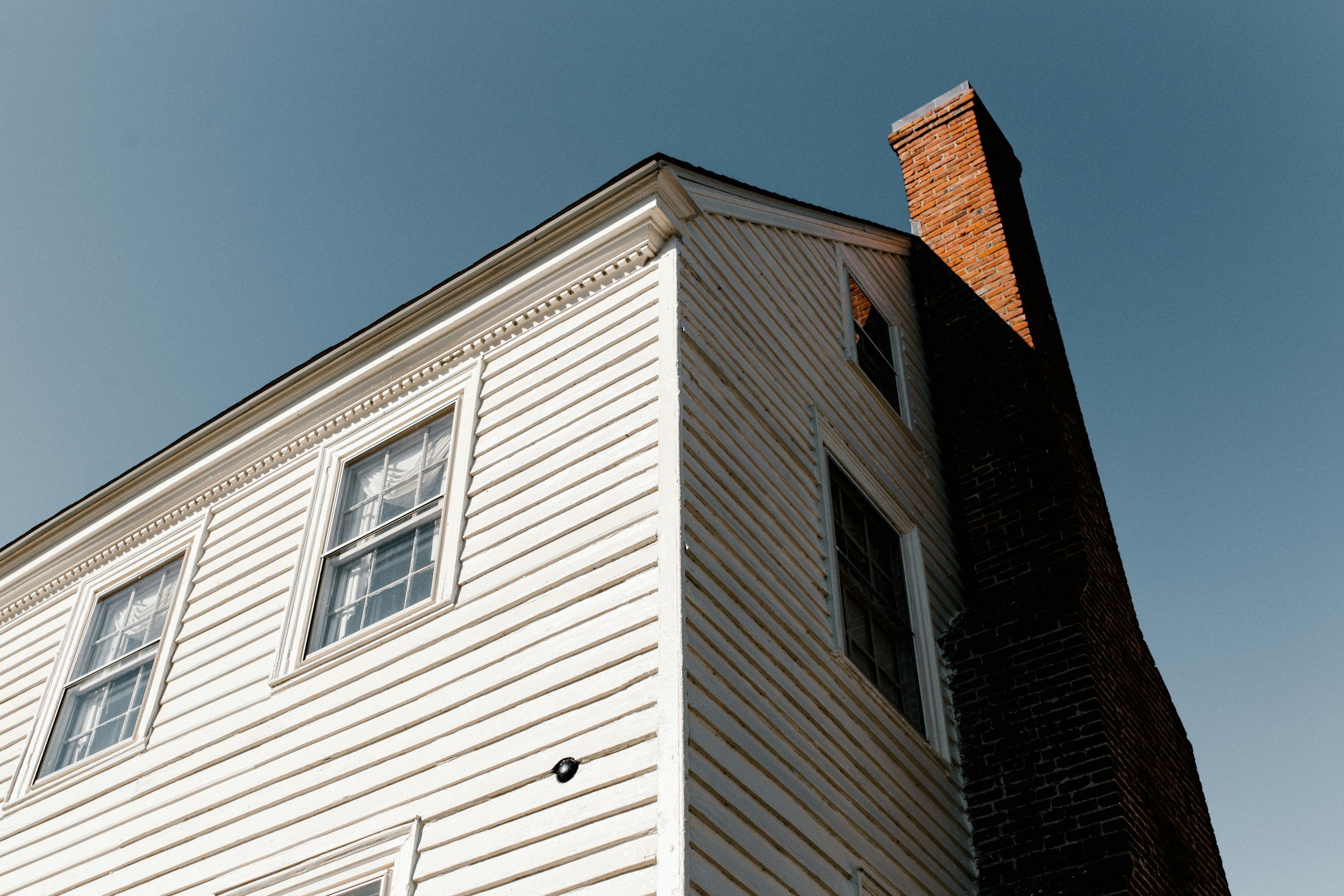 Close-up of the corner of a white, wooden, two-story house with a brick chimney and three windows, under a clear blue sky.