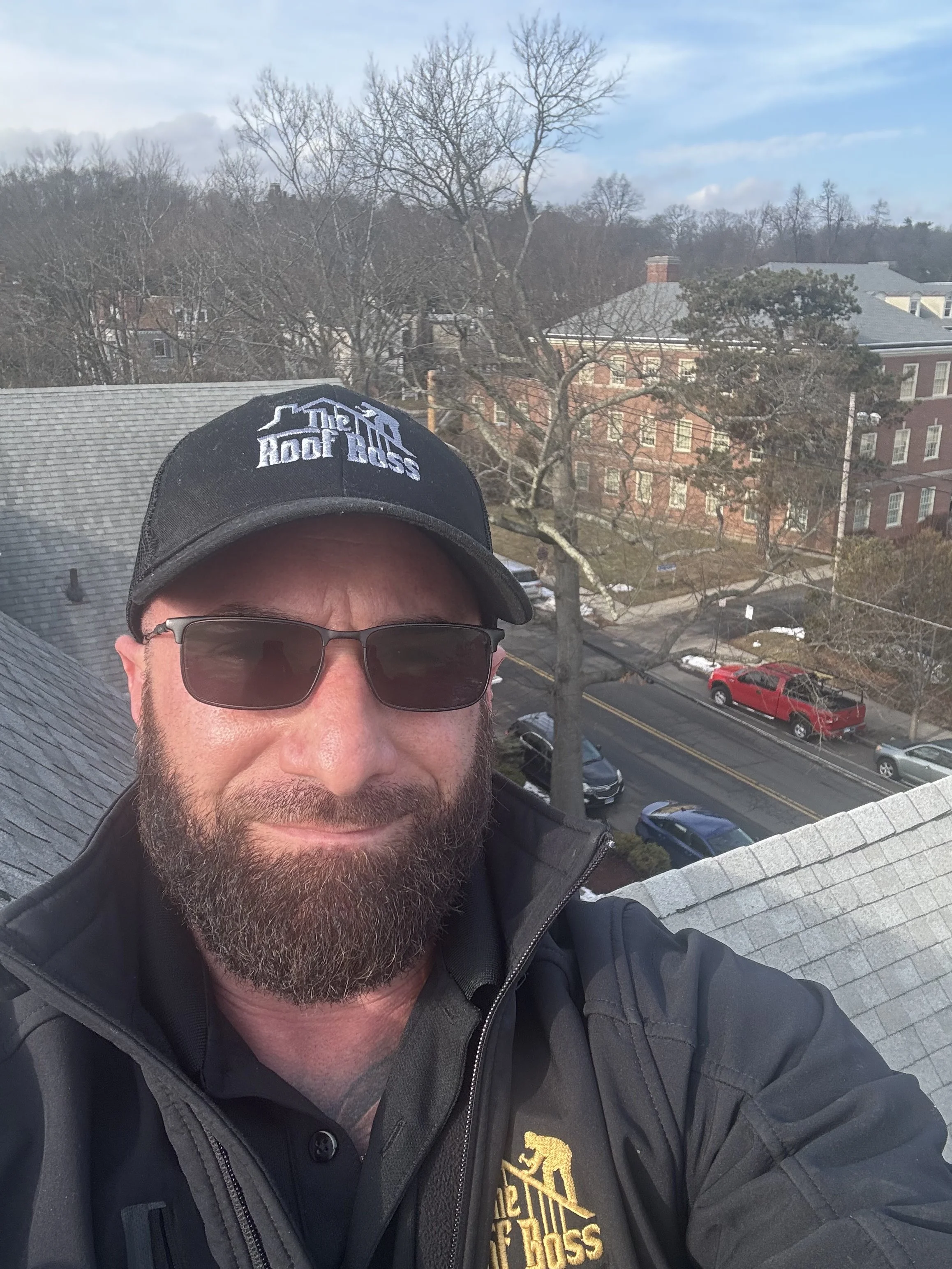 A man with a beard wearing sunglasses, a black cap with 'Man Roof Boss' embroidered on it, and a black jacket with the same logo, standing on a rooftop with a view of a street and buildings in the background.