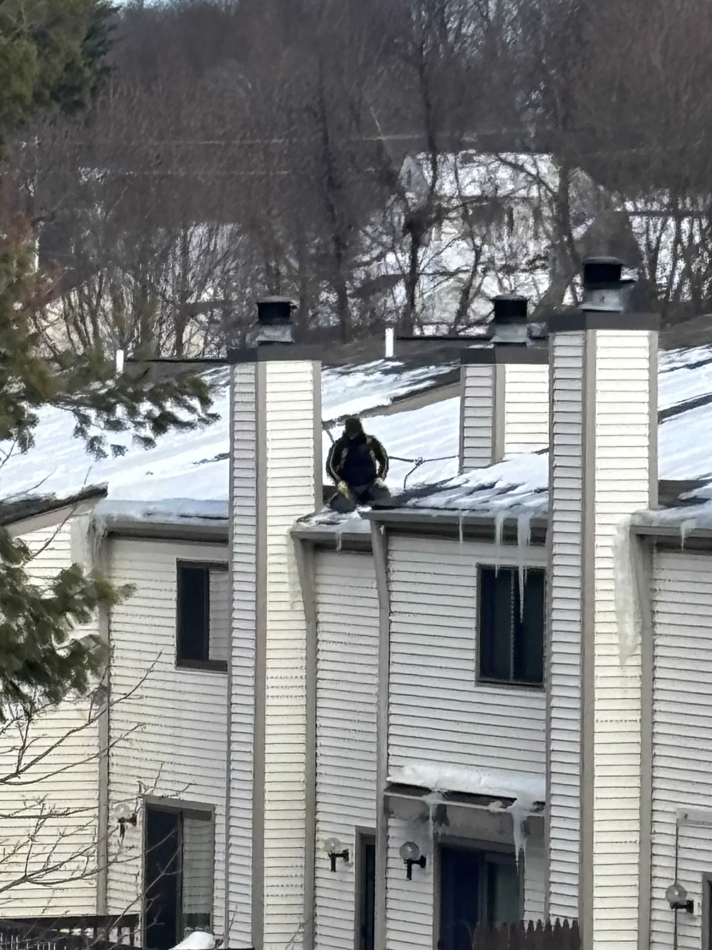 A person on a ladder cleaning or inspecting a snow-covered roof of a white house during winter.