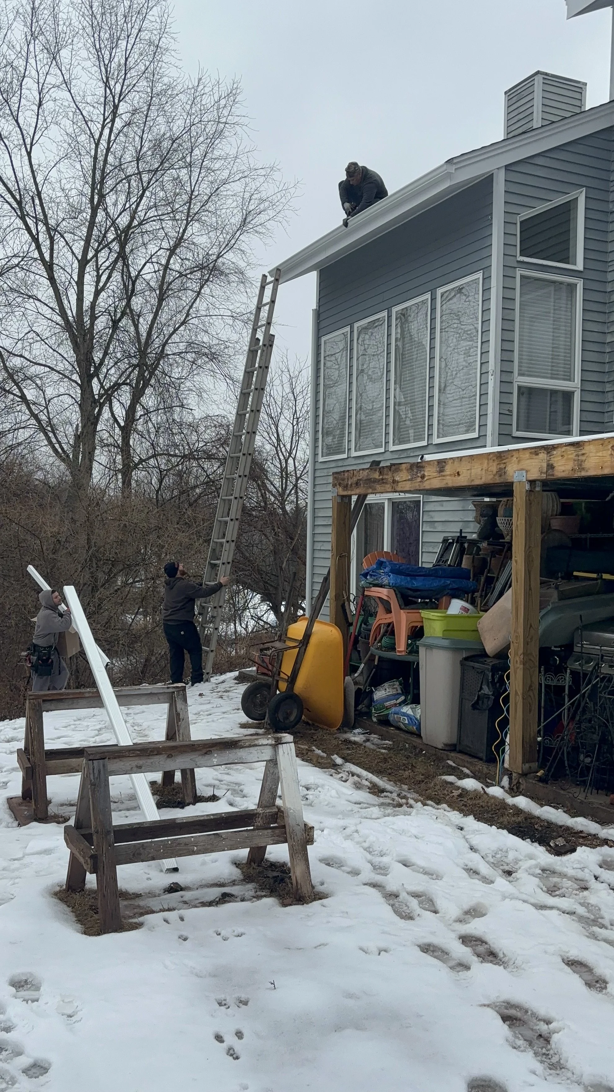 People working on installing or repairing the gutter of a house with a ladder, with snow on the ground.