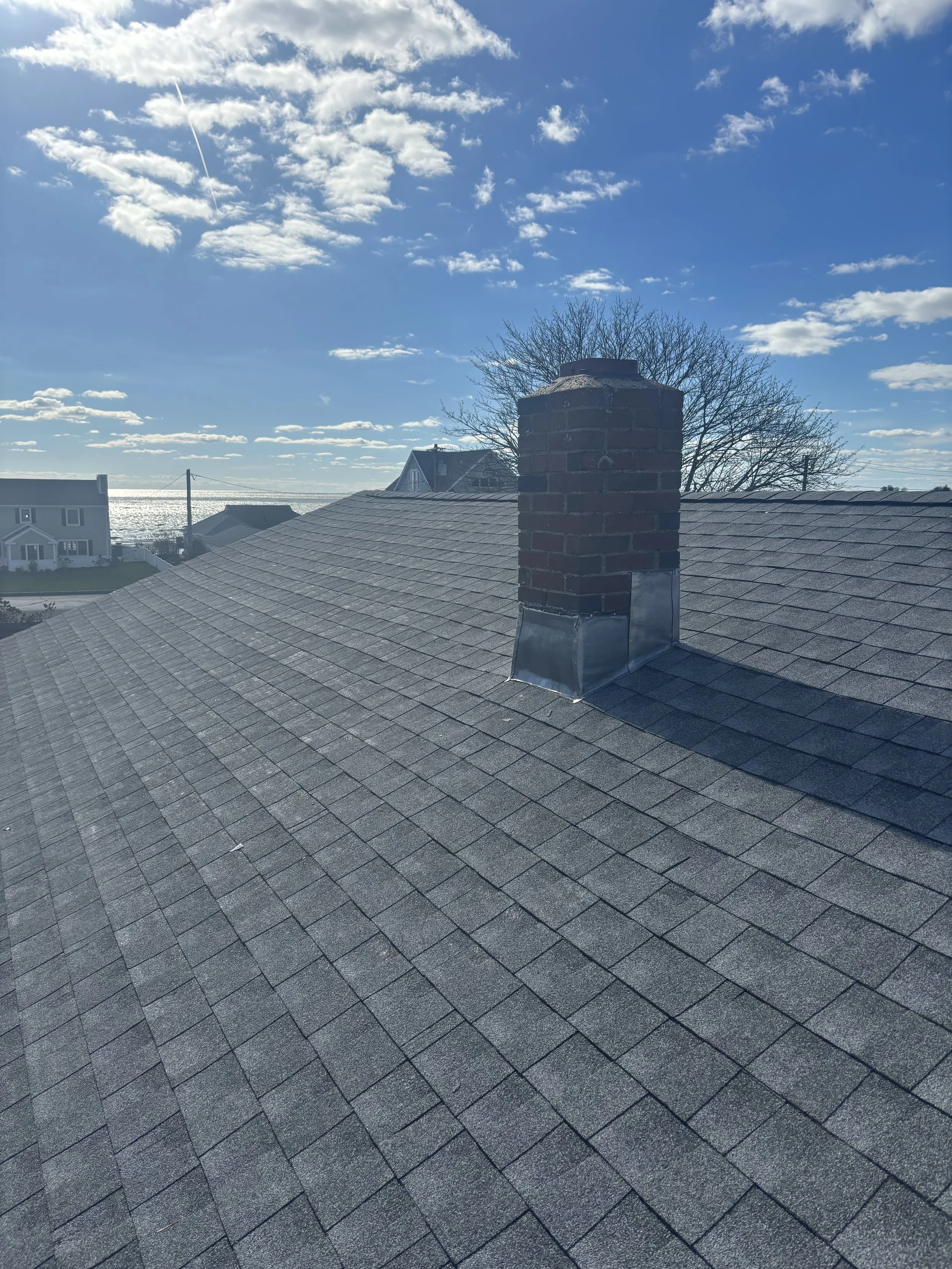 View of a roof with gray shingles and a brick chimney under a blue sky with clouds.