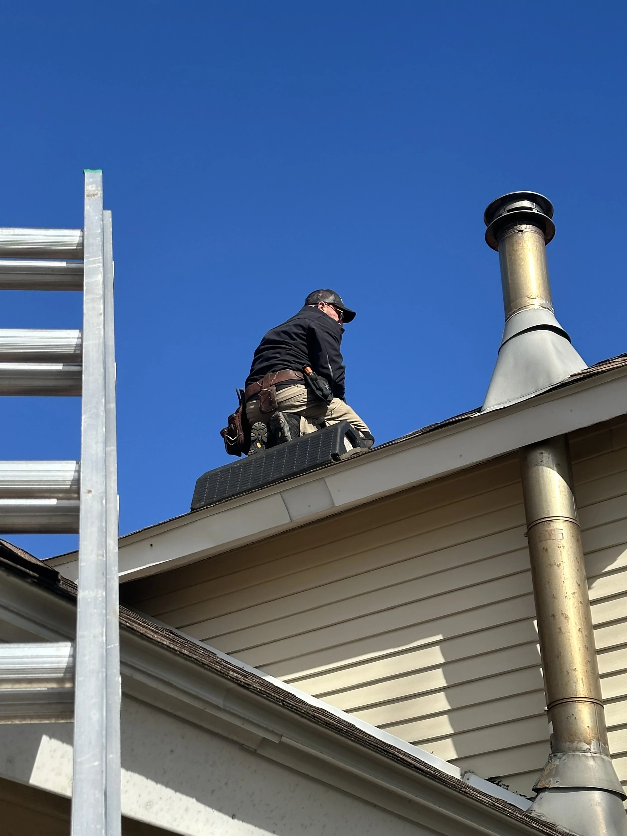 A worker on a house roof, wearing work gear, kneeling near a chimney, with a ladder visible on the left side and a clear blue sky in the background.