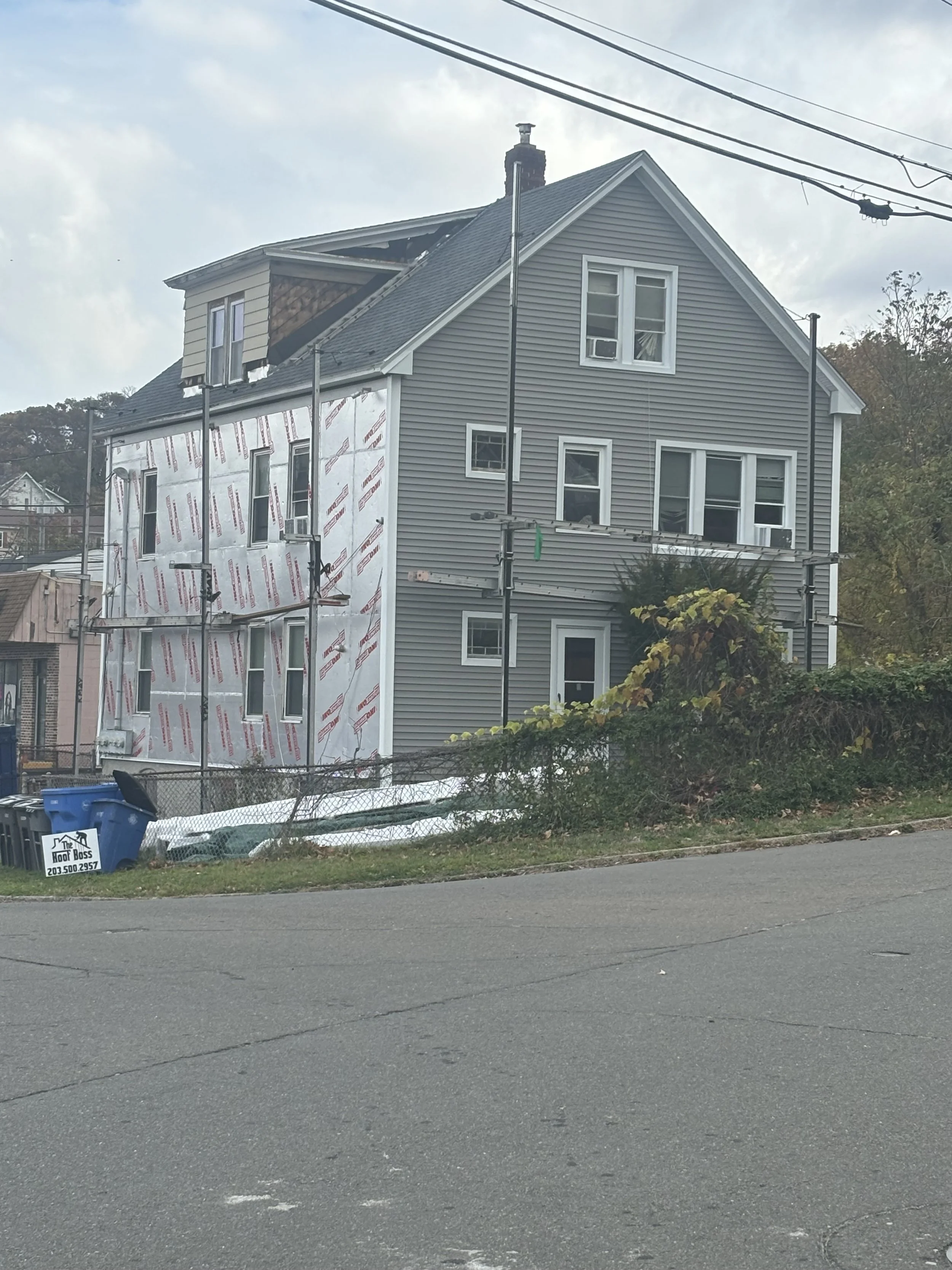 Side of a house under renovation with grey siding, partially covered in white insulation wrap, and scaffolding set up around it. The house is on a corner lot with a small yard and a chain-link fence. There are power lines overhead.
