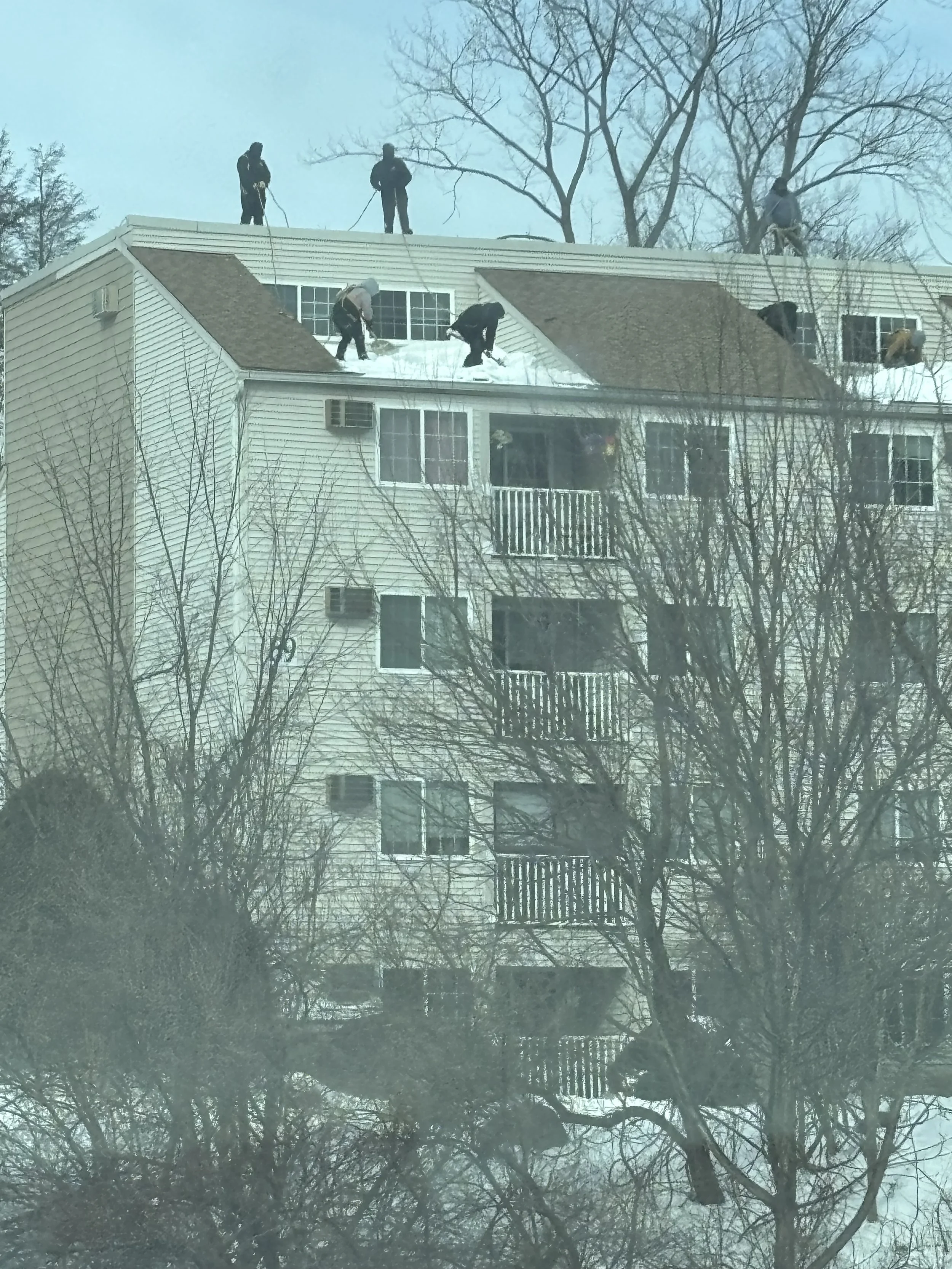 Six people working on snow removal on the roof of a multi-story beige apartment building during winter, with leafless trees in the foreground.