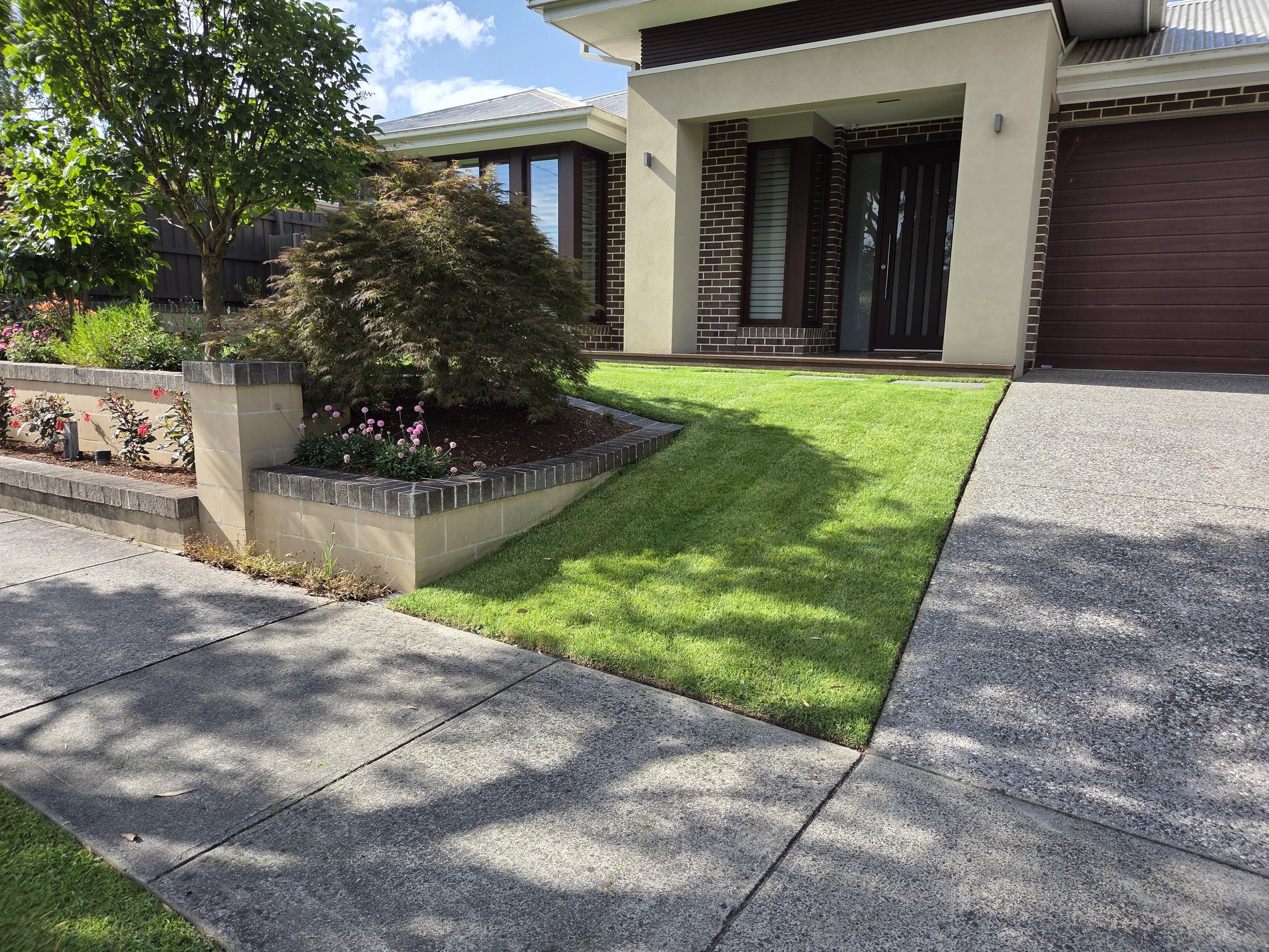 Front yard with green grass, a small garden bed with trees and flowers, and a concrete driveway leading to a house with a brick and stucco exterior.