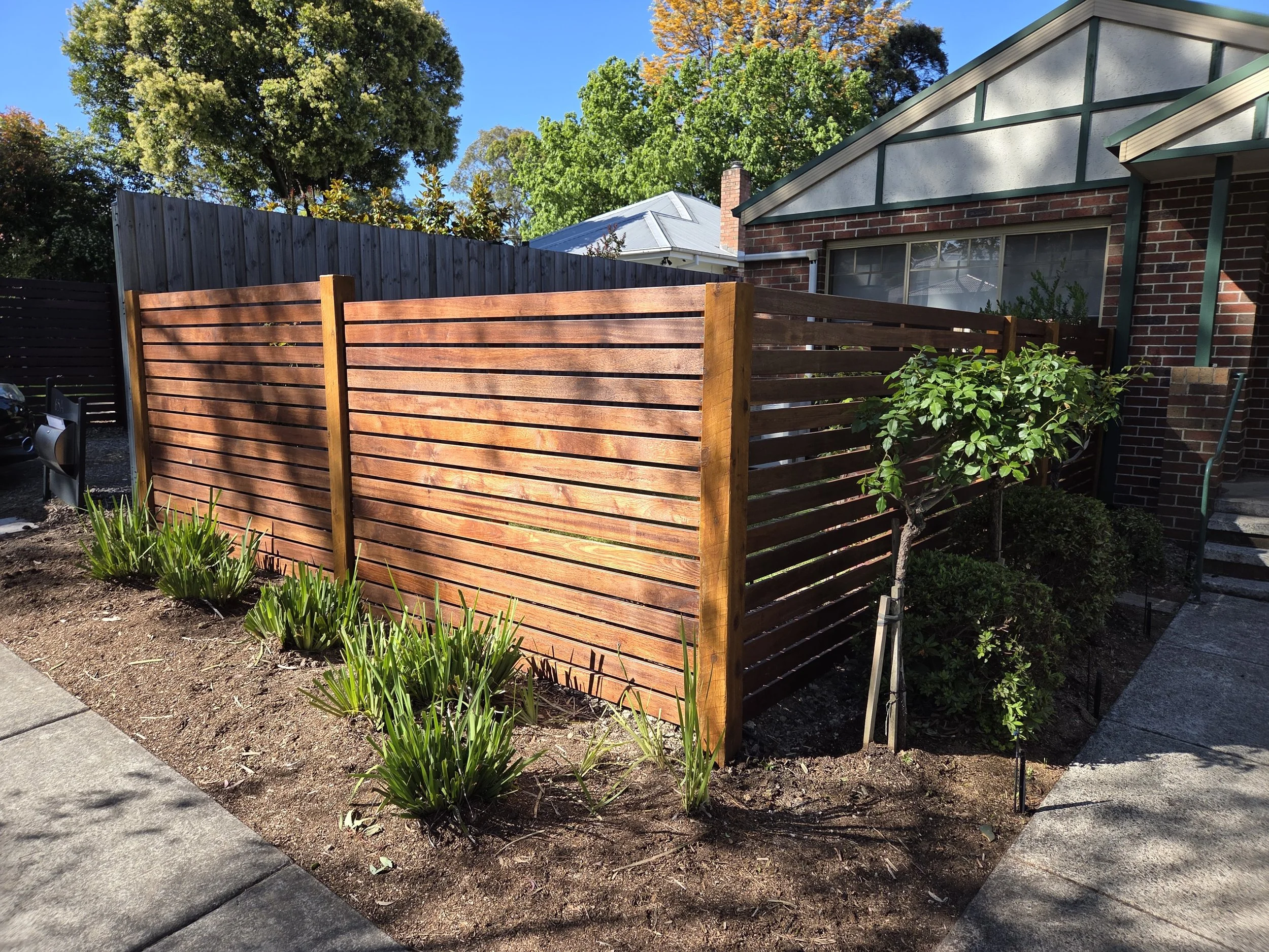 A wooden privacy fence surrounds an outdoor area in front of a house with brick walls and steps. There are small bushes and plants along the base of the fence and a tree nearby. The sky is clear and blue.