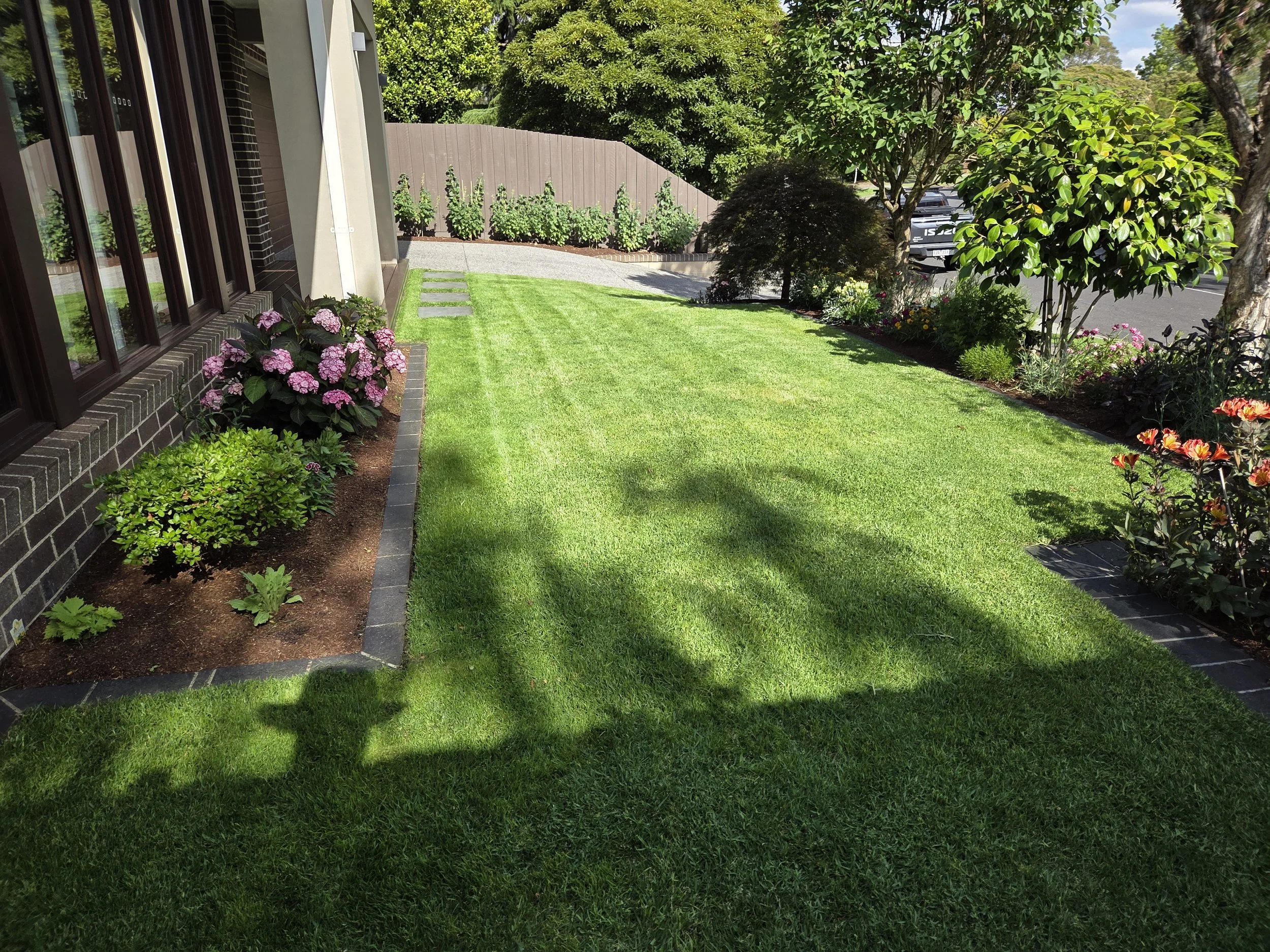 A well-maintained front yard with green grass, flower beds along the house and front fence, bushes, and trees, with parked cars visible on the street in the background.