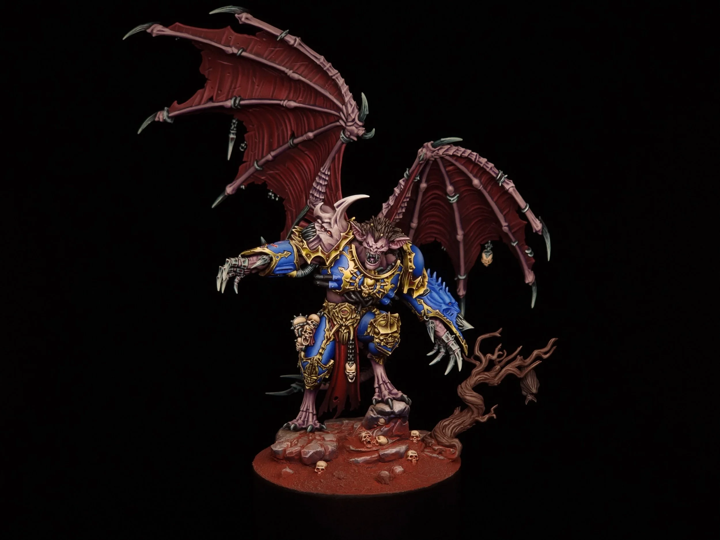A detailed miniature model of a dragon-like creature with bat wings, armored in blue and gold, standing on a rocky base with skull decorations and a twisted tree, set against a black background.