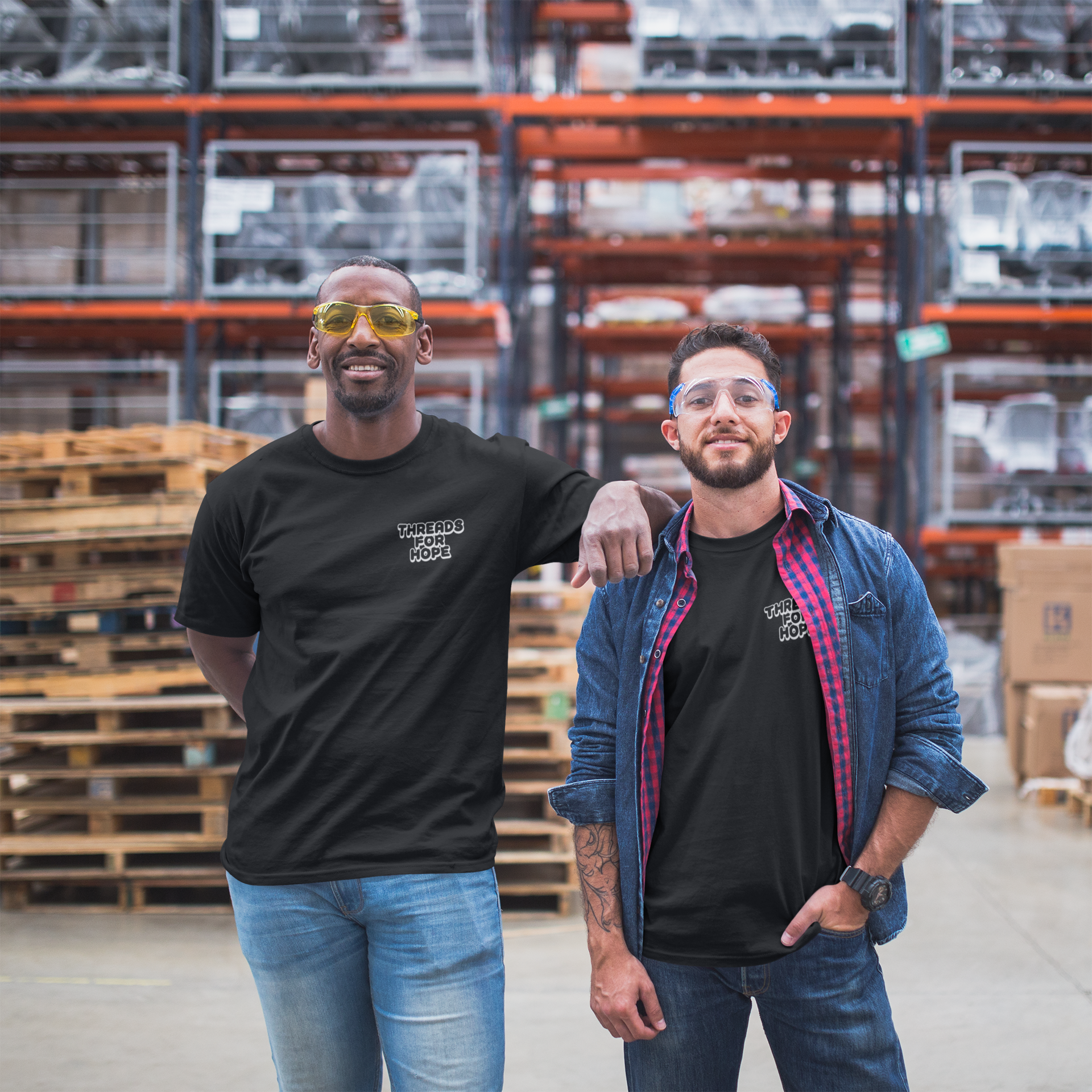 Two men standing in a warehouse, both wearing glasses and black t-shirts with 'Threads for Hope' logo, one with arm resting on the other's shoulder, surrounded by stacks of wooden pallets and industrial shelving.