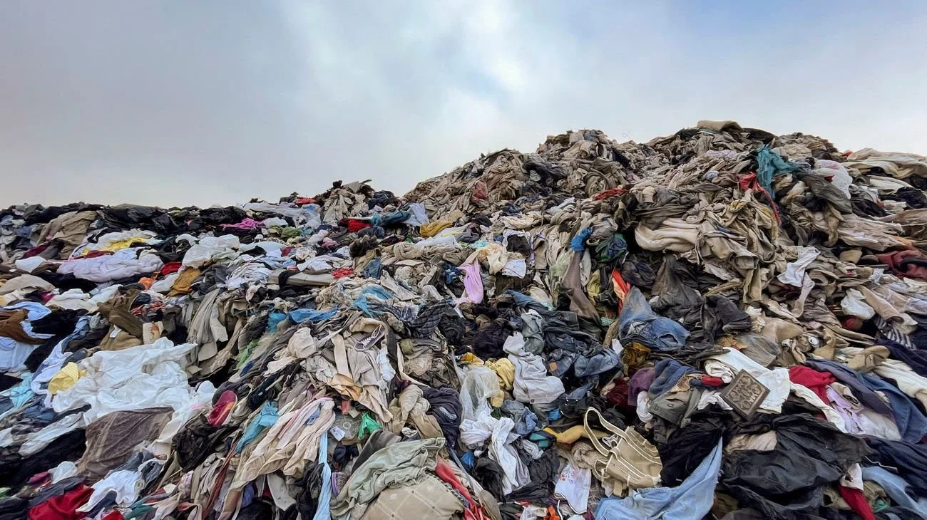 A large pile of discarded clothes and fabric waste outdoors against a cloudy sky.