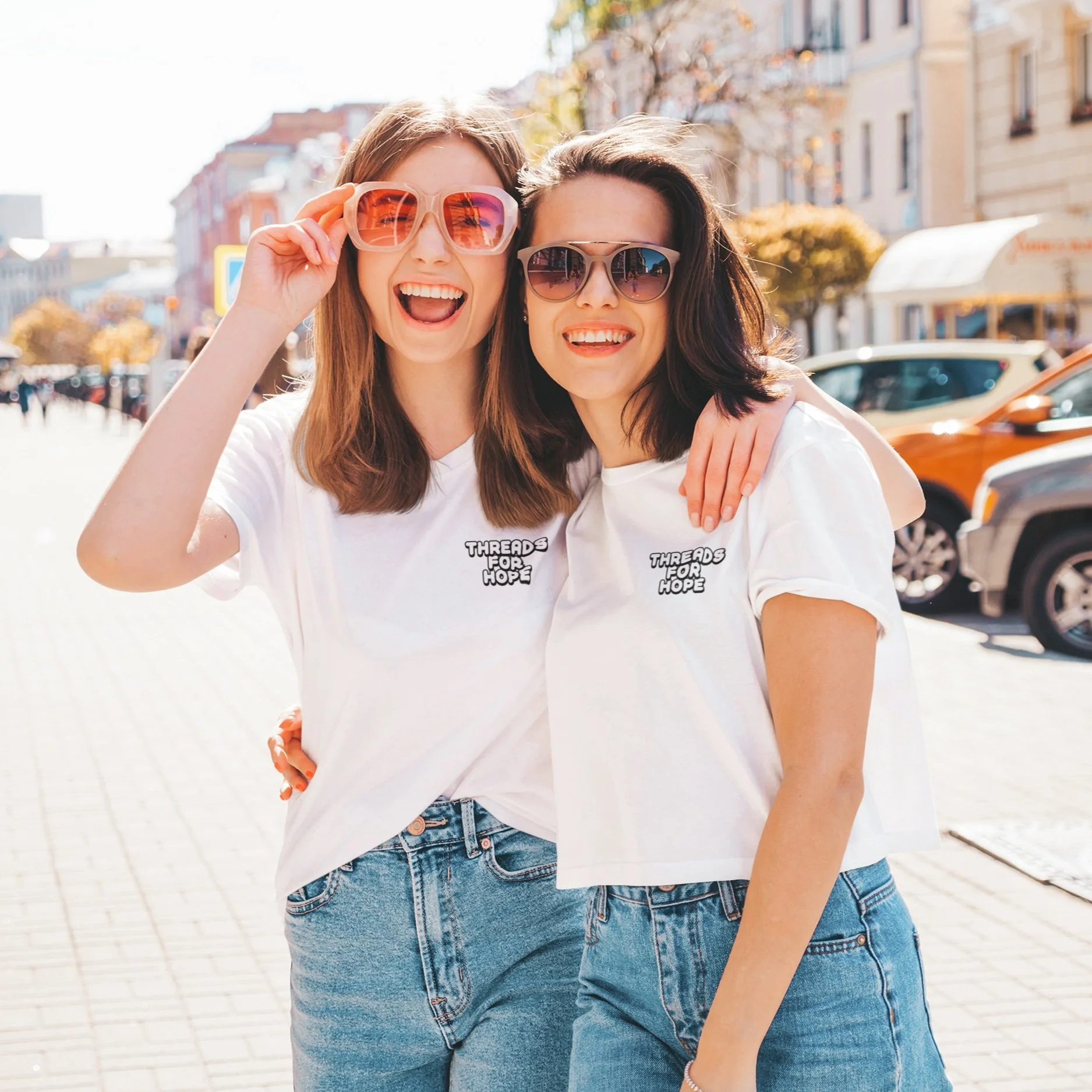 Two young women wearing sunglasses and white T-shirts with 'Threads for Hope' logo, smiling and posing outdoors in a city street on sunny day.
