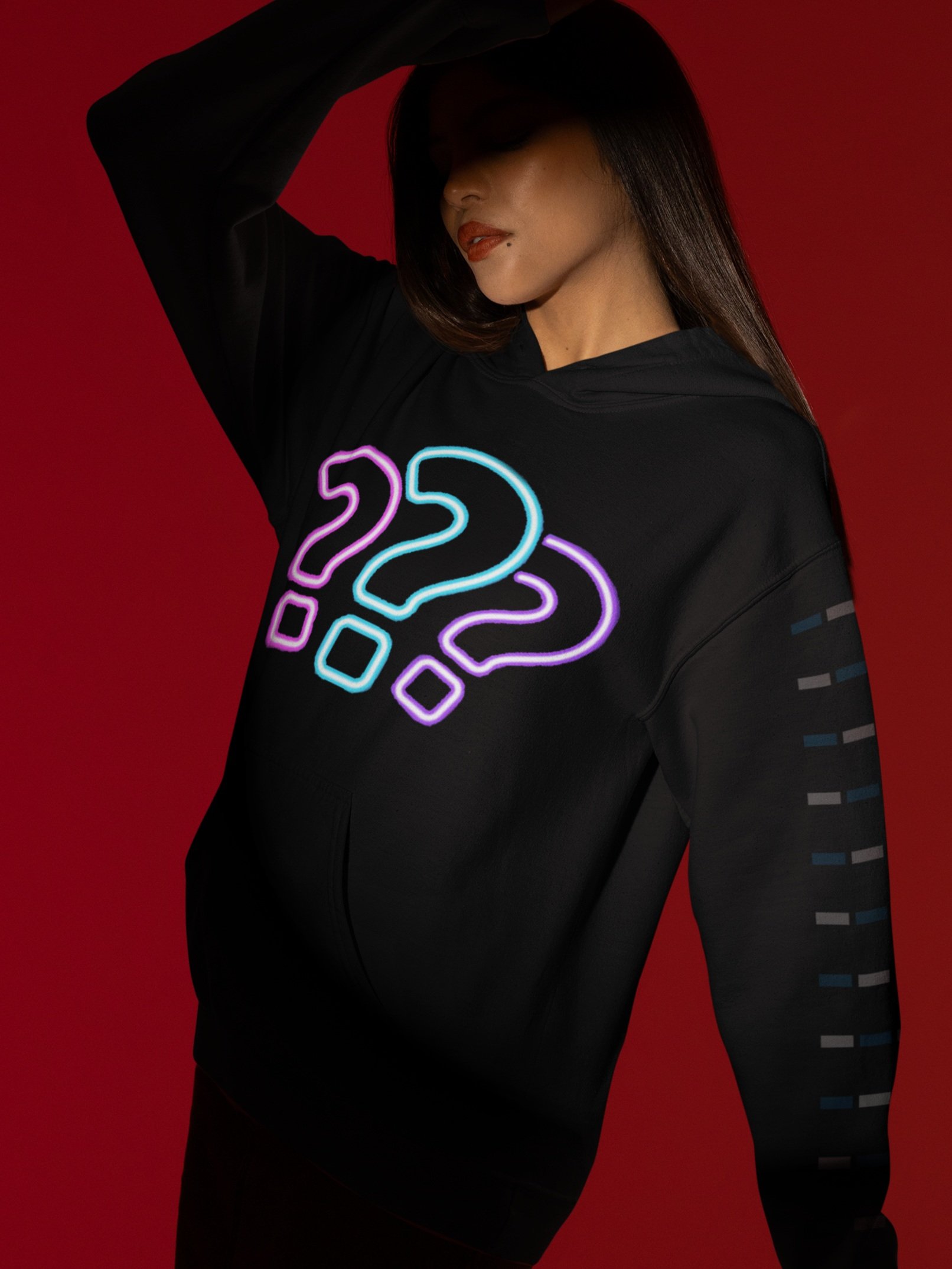 A woman with long dark hair wearing a black hoodie with neon-colored question marks on the front, posing against a red background.