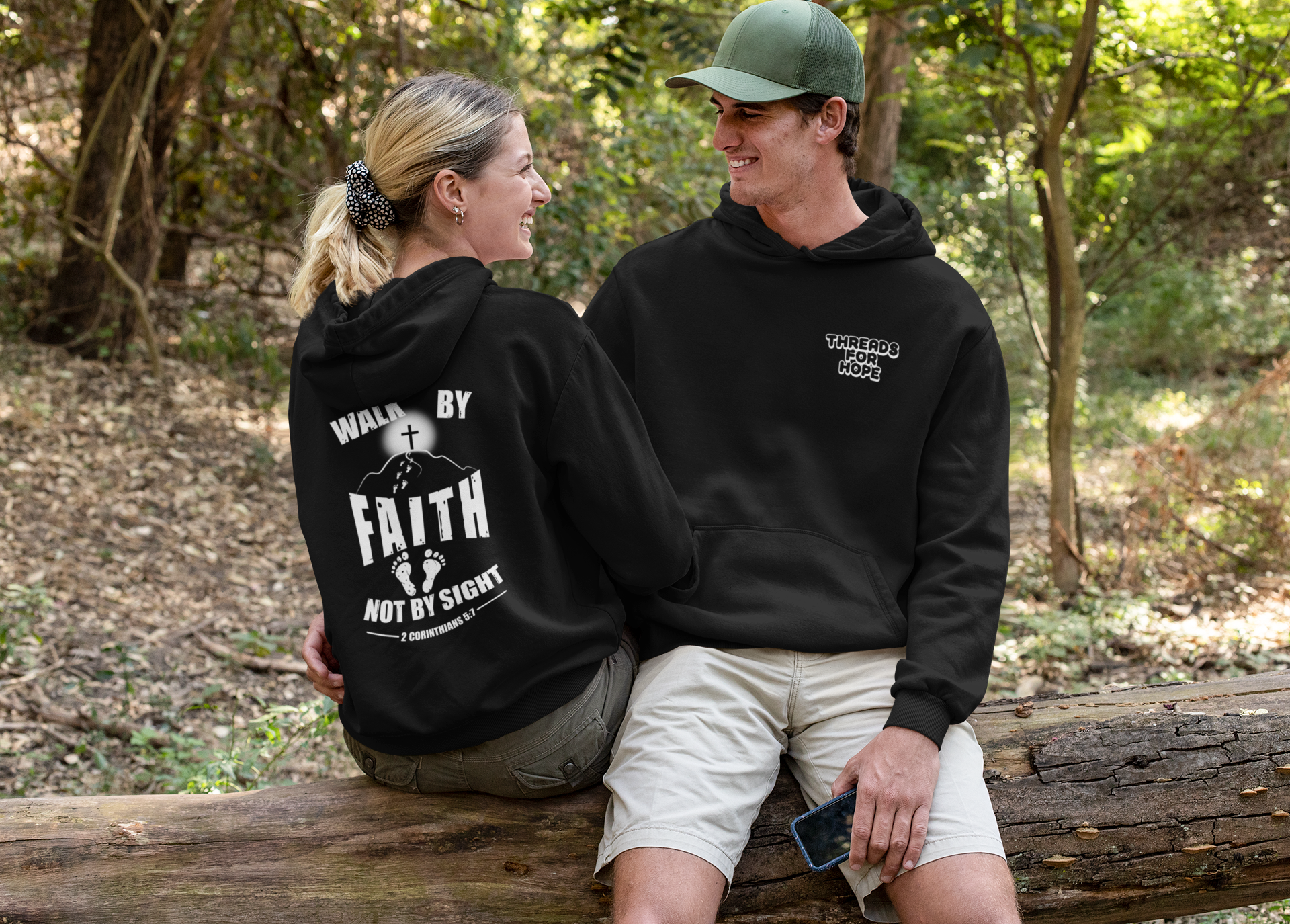 A man and a woman sit on a log in a wooded area, smiling and looking at each other. Both are wearing black sweatshirts with white text and designs. The woman's sweatshirt reads "WALK BY FAITH NOT BY SIGHT" with a cross and feet graphic, and the man's sweatshirt reads "THREADS FOR HOPE."