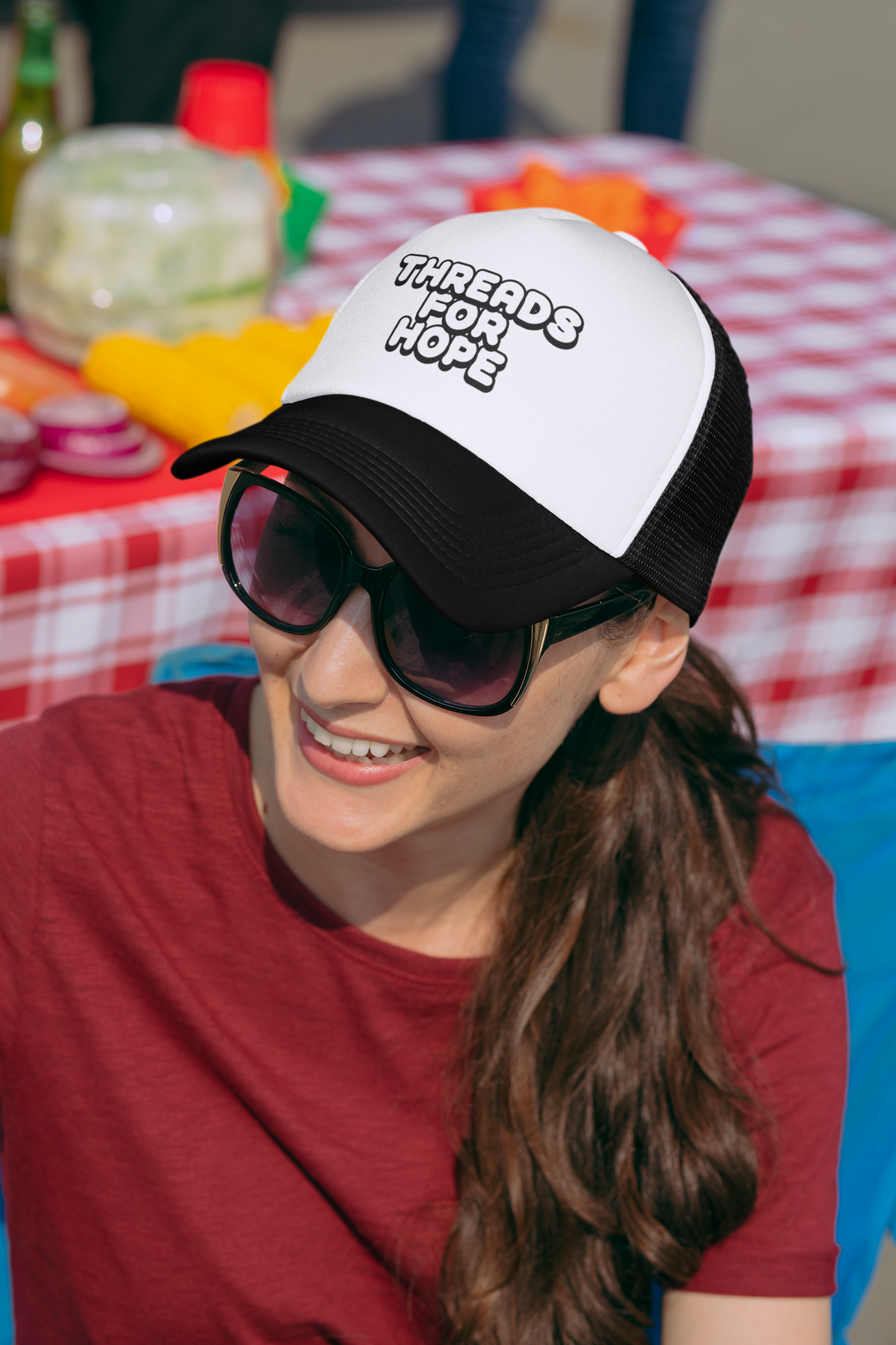 Smiling woman wearing sunglasses and a trucker hat with the phrase "Threads for Hope" at an outdoor gathering with a red and white checkered tablecloth and food in the background.