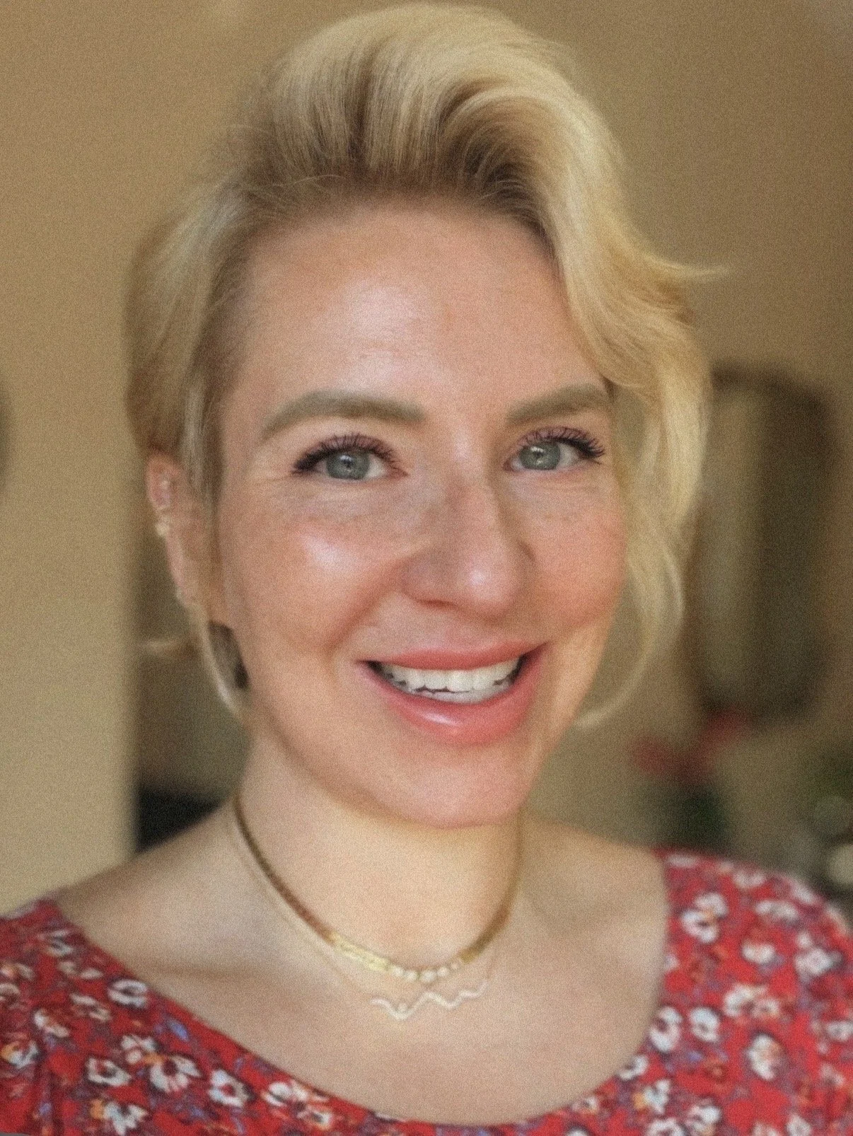 Close-up of a smiling woman with short blonde hair, blue eyes, and light skin, wearing a red floral top and layered necklaces.