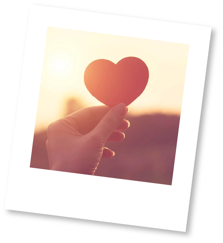 A hand holding a paper heart in front of a sunset.